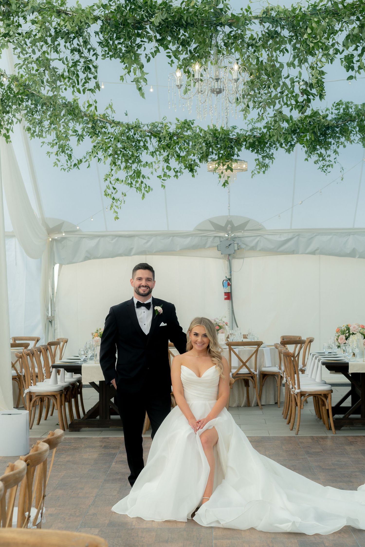 Bride and groom pose elegantly inside a tented reception venue adorned with lush greenery chandeliers.