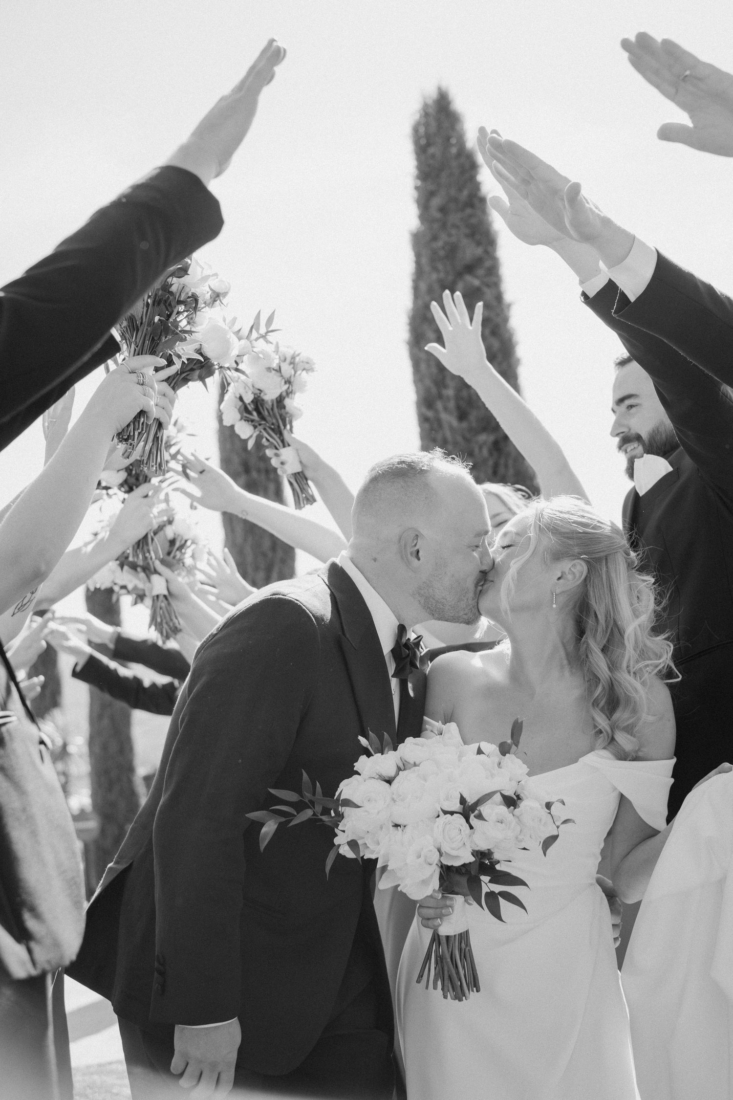 Black and white photo of newlyweds sharing first kiss under arch of raised hands from wedding party.