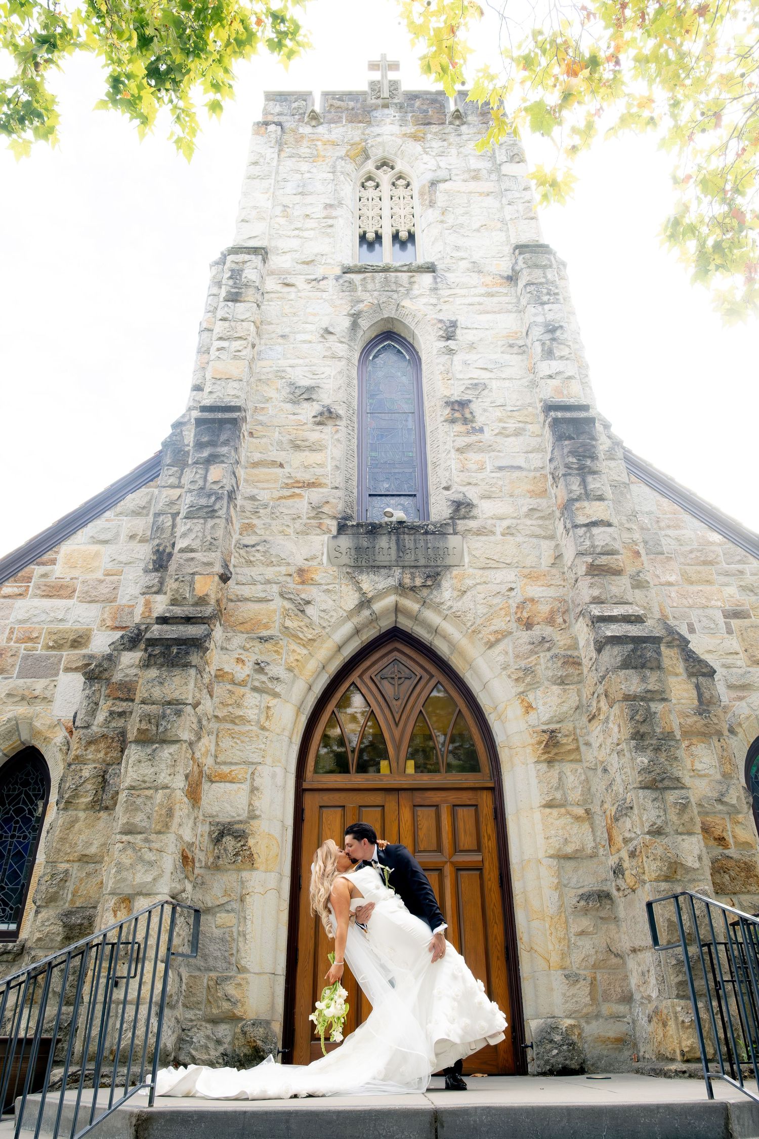 Bride and groom sharing romantic dip kiss outside stone Gothic church after wedding ceremony.