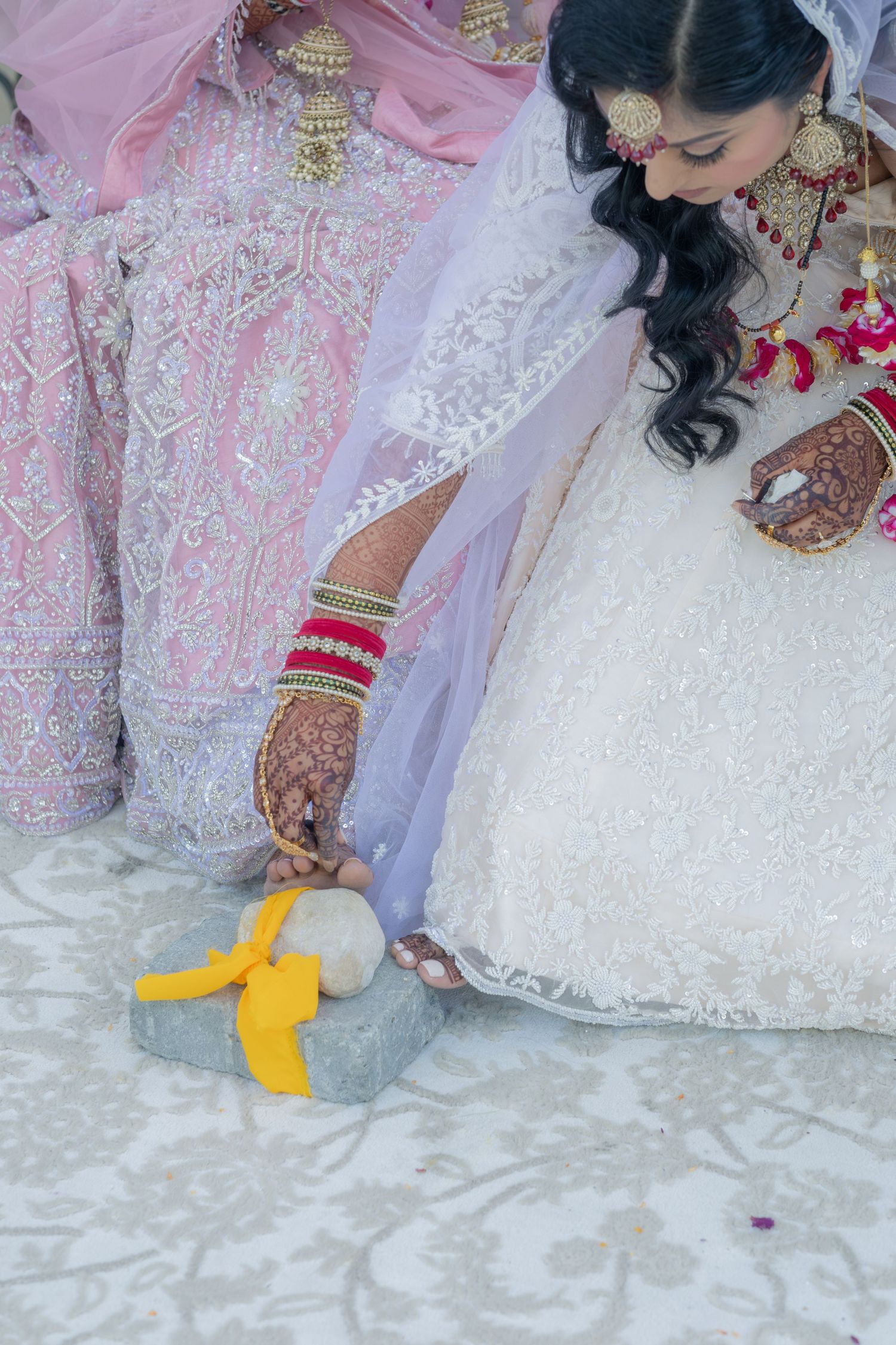 Indian bride in white lehenga with mehndi hands performing wedding ritual on stone step with yellow cloth.