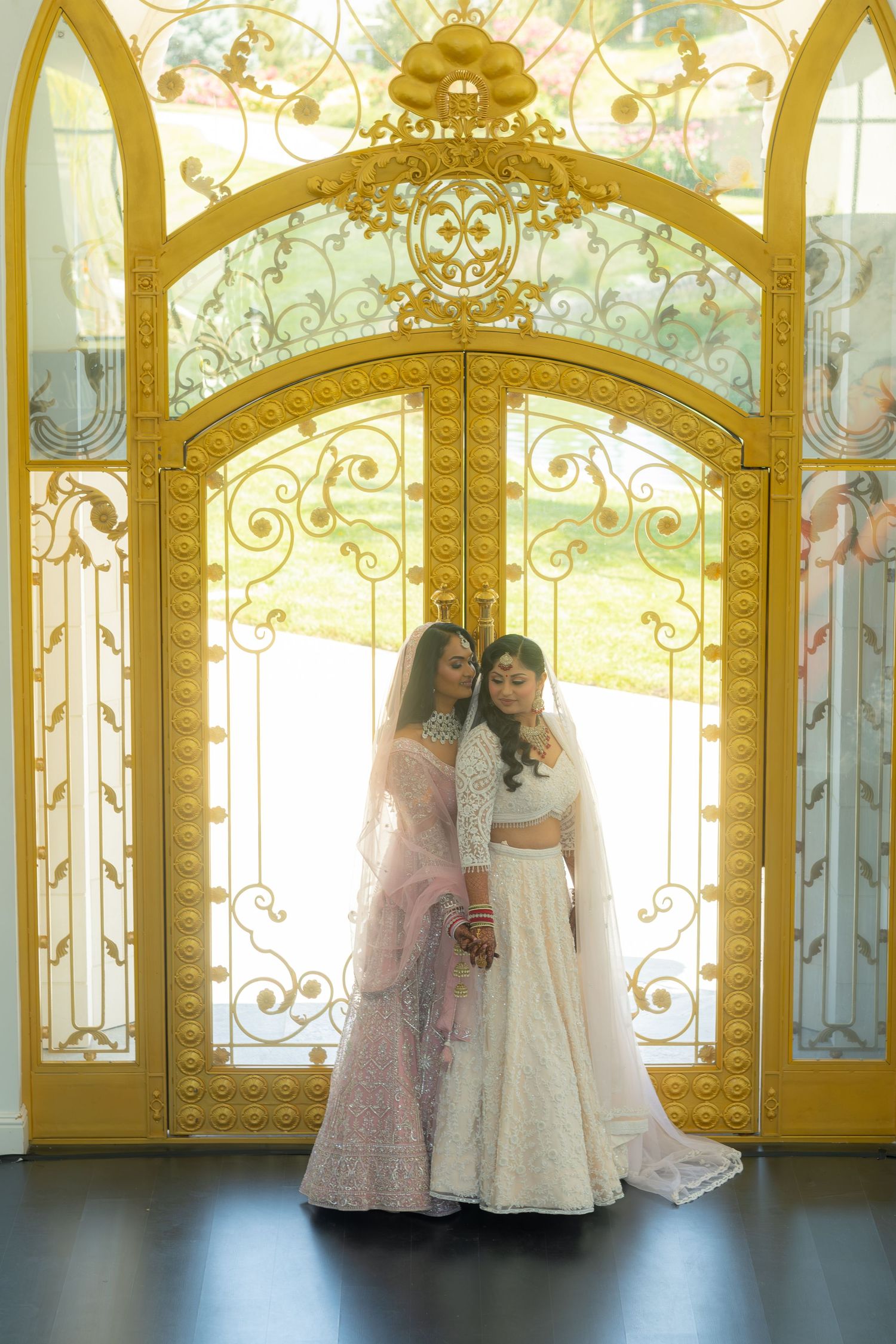 Two women in white and pink Indian bridal outfits pose before ornate golden arched doors.