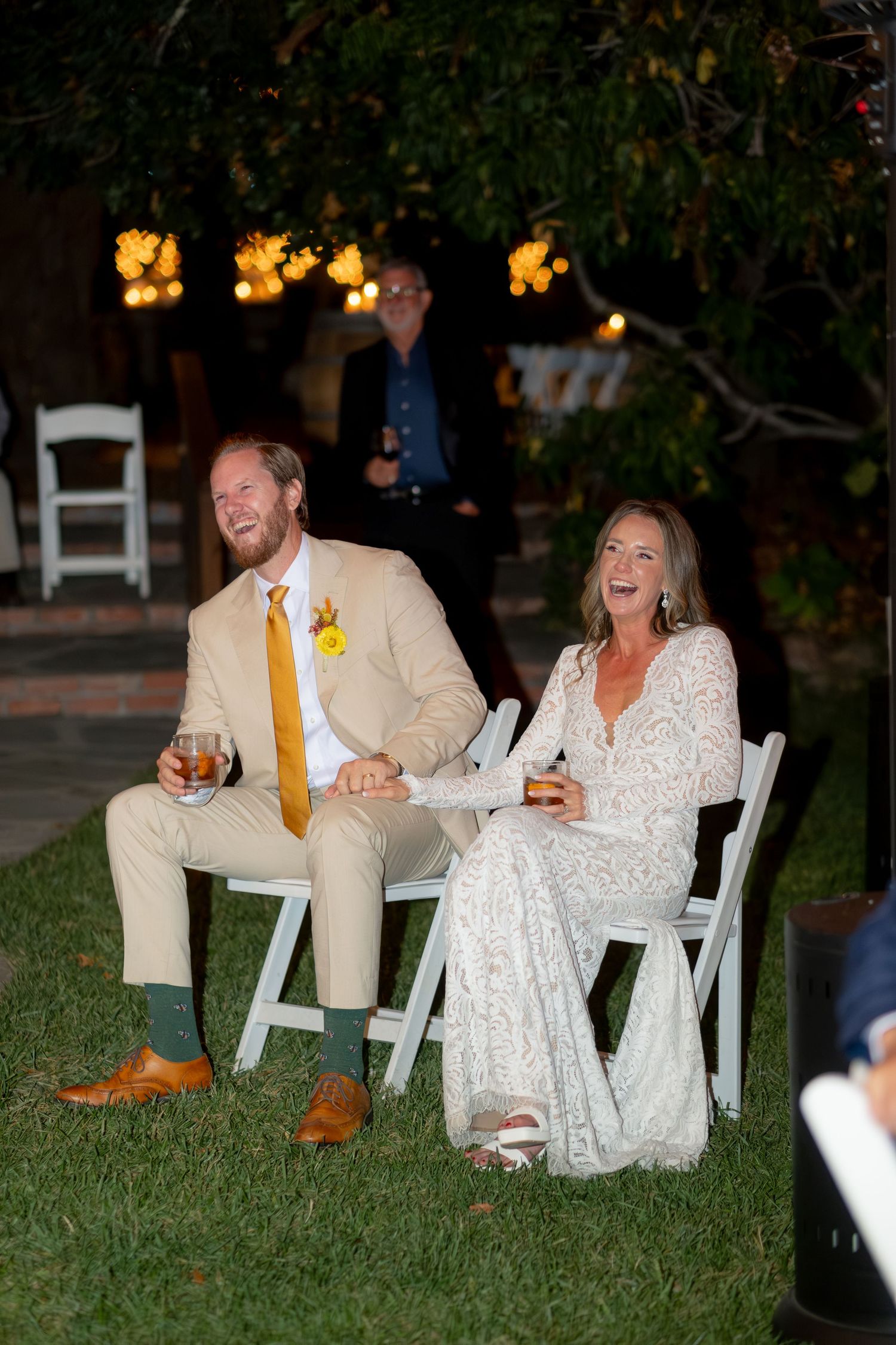 Bride and groom laughing together seated in white chairs at outdoor evening wedding reception.