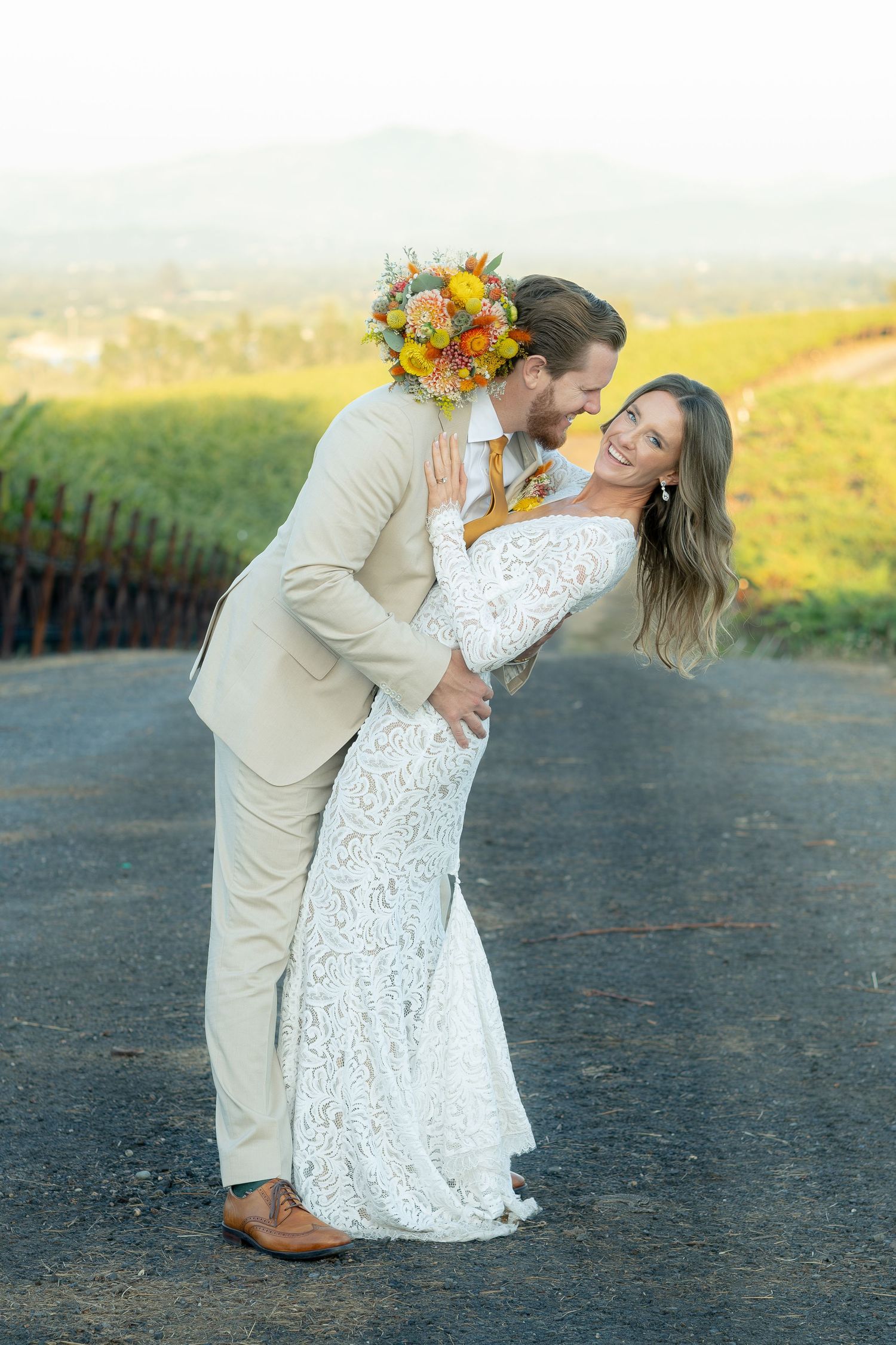 Groom dips smiling bride holding colorful bouquet on a road with vineyard and mountain backdrop.