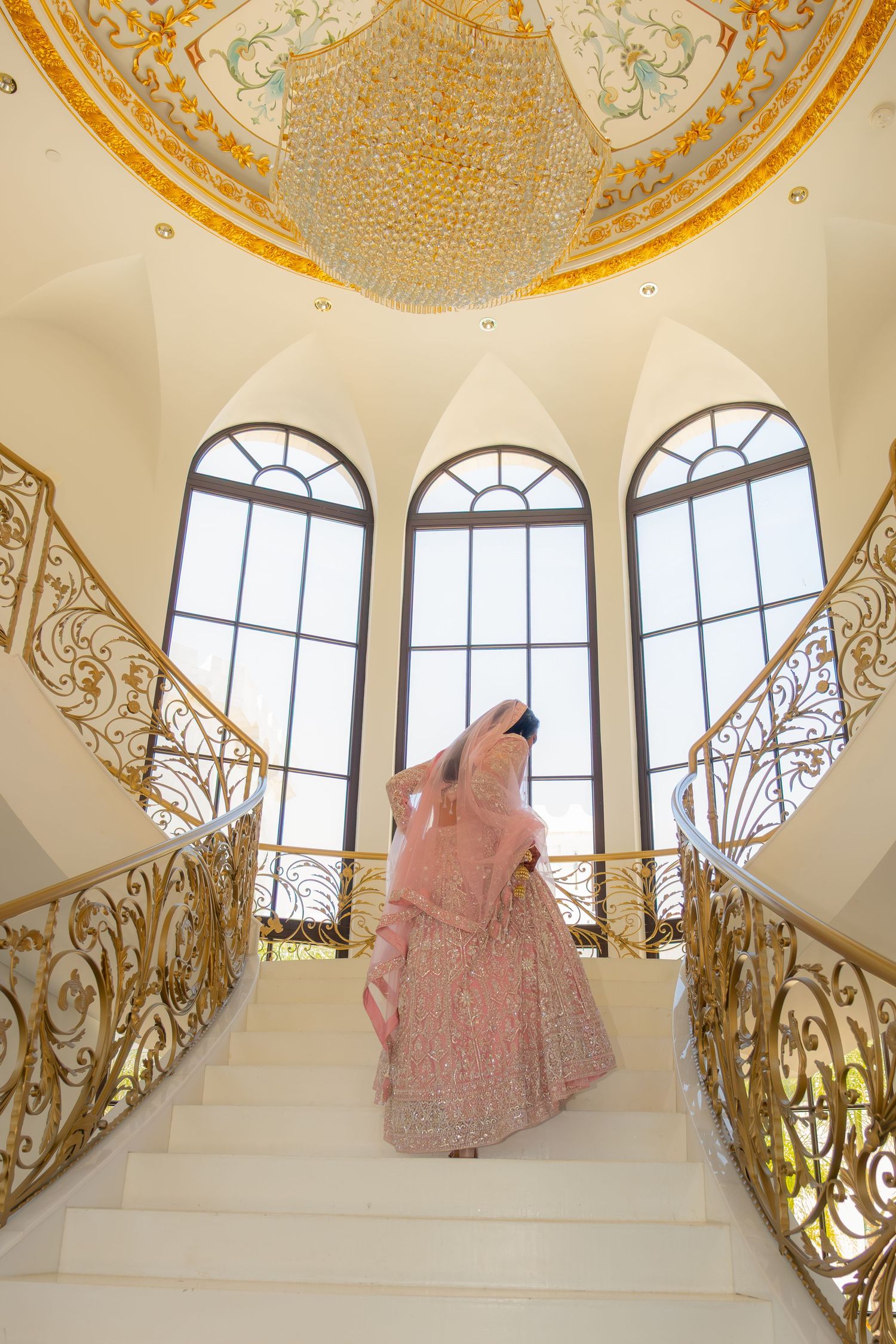 Indian bride in pink lehenga ascending grand staircase under ornate crystal chandelier.