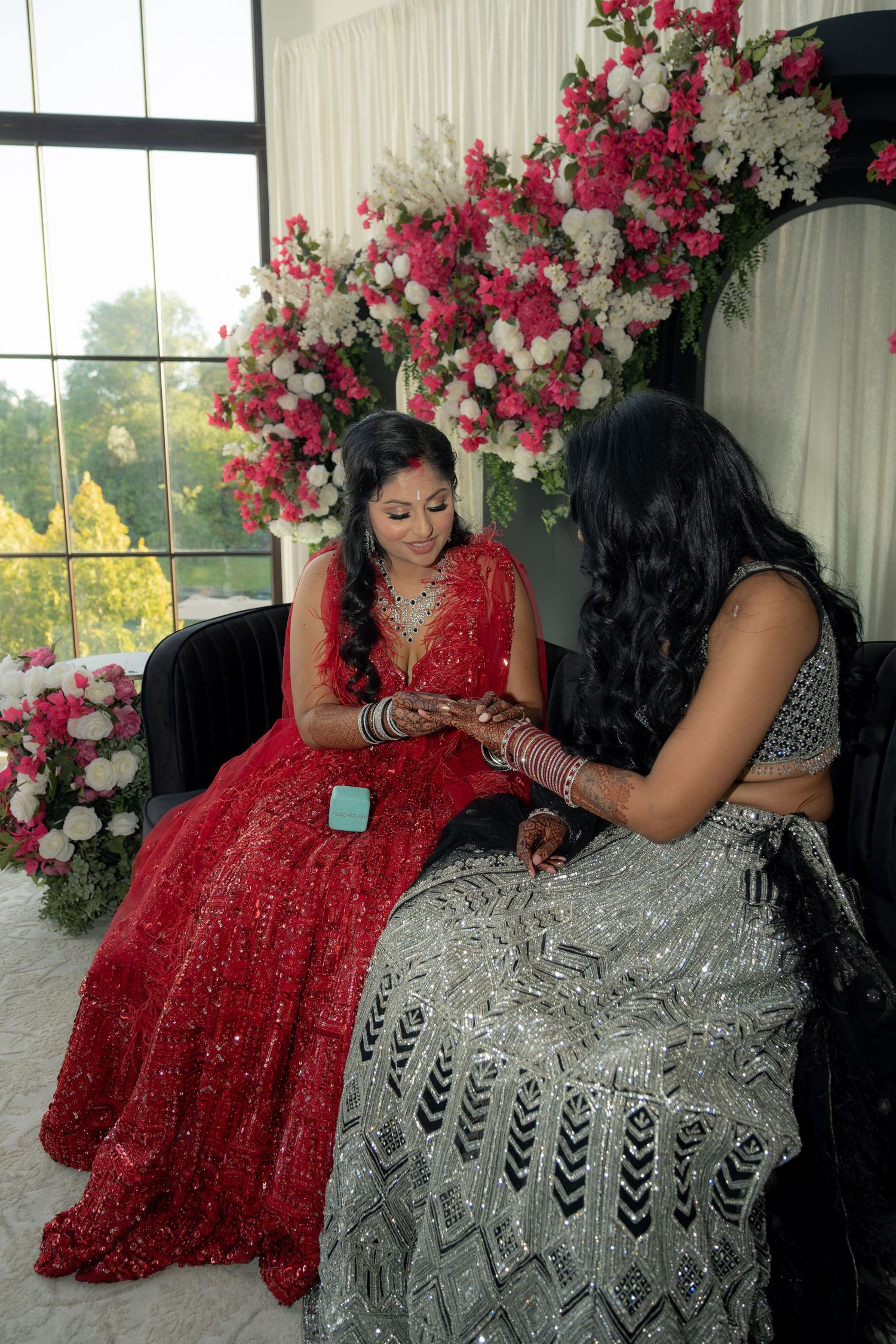 Two women in red and silver Indian bridal attire sit together examining jewelry amid pink and white floral decor.
