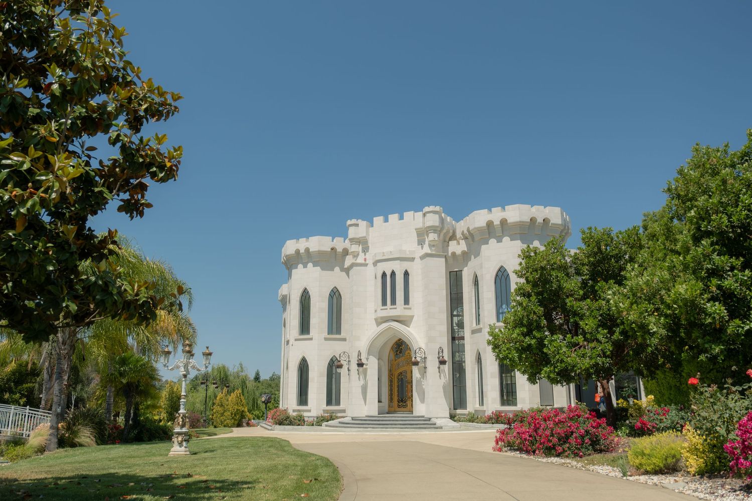 White castle-style building with Gothic arched windows, surrounded by lush trees and colorful gardens under a clear blue sky.