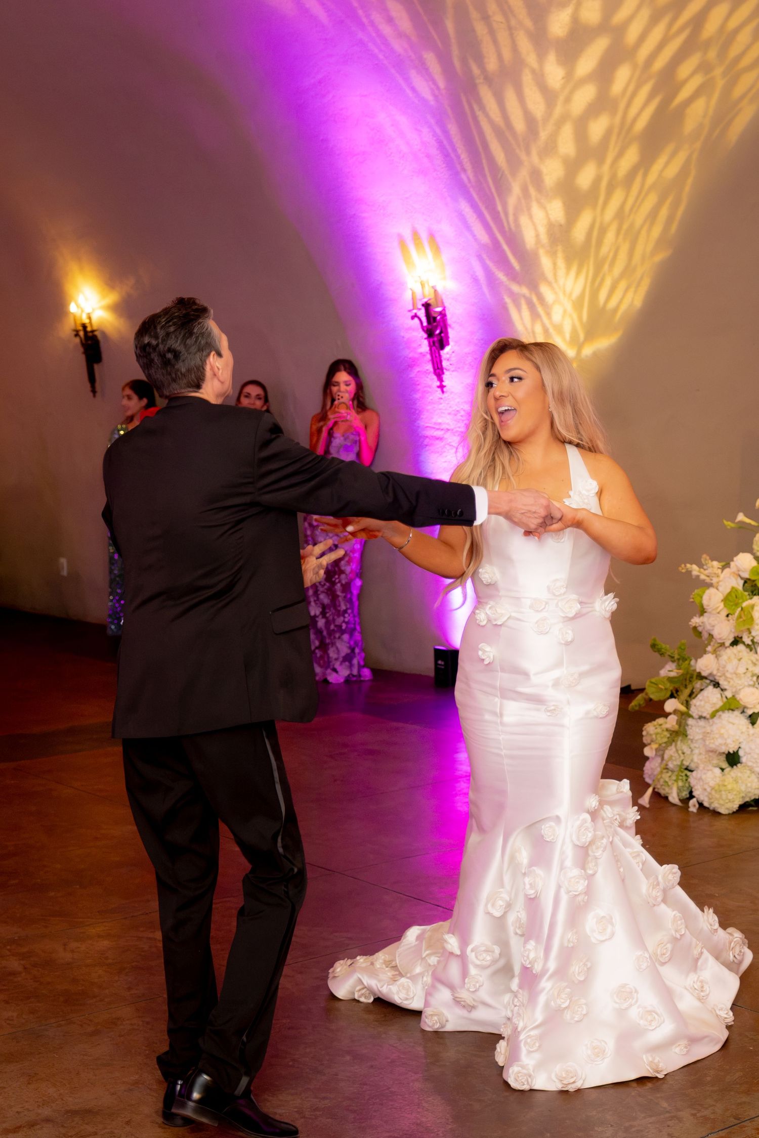 Bride in floral mermaid gown dances joyfully with groom under purple uplighting at wedding reception.