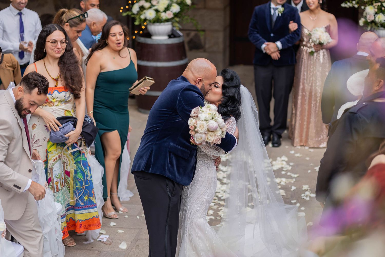 Groom in navy velvet suit kissing bride in white gown as guests celebrate along the aisle.