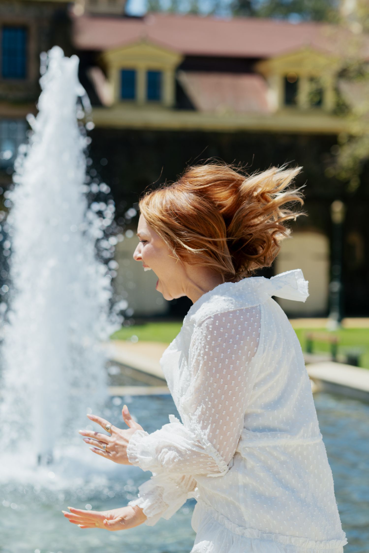Joyful woman in white lace dress standing near a fountain with hair blowing in the breeze.