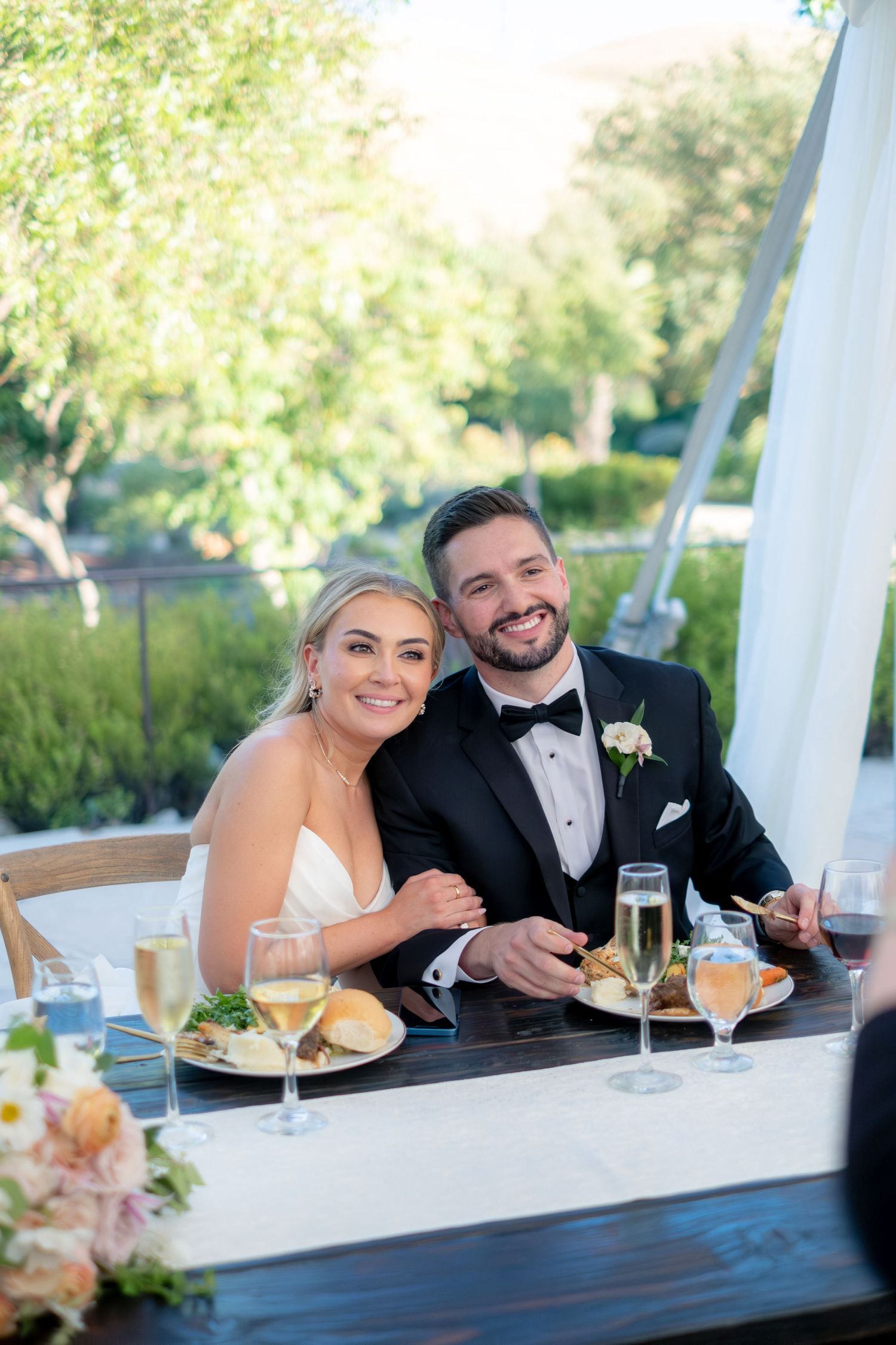 Smiling bride and groom seated at sweetheart table during outdoor wedding reception with champagne glasses.
