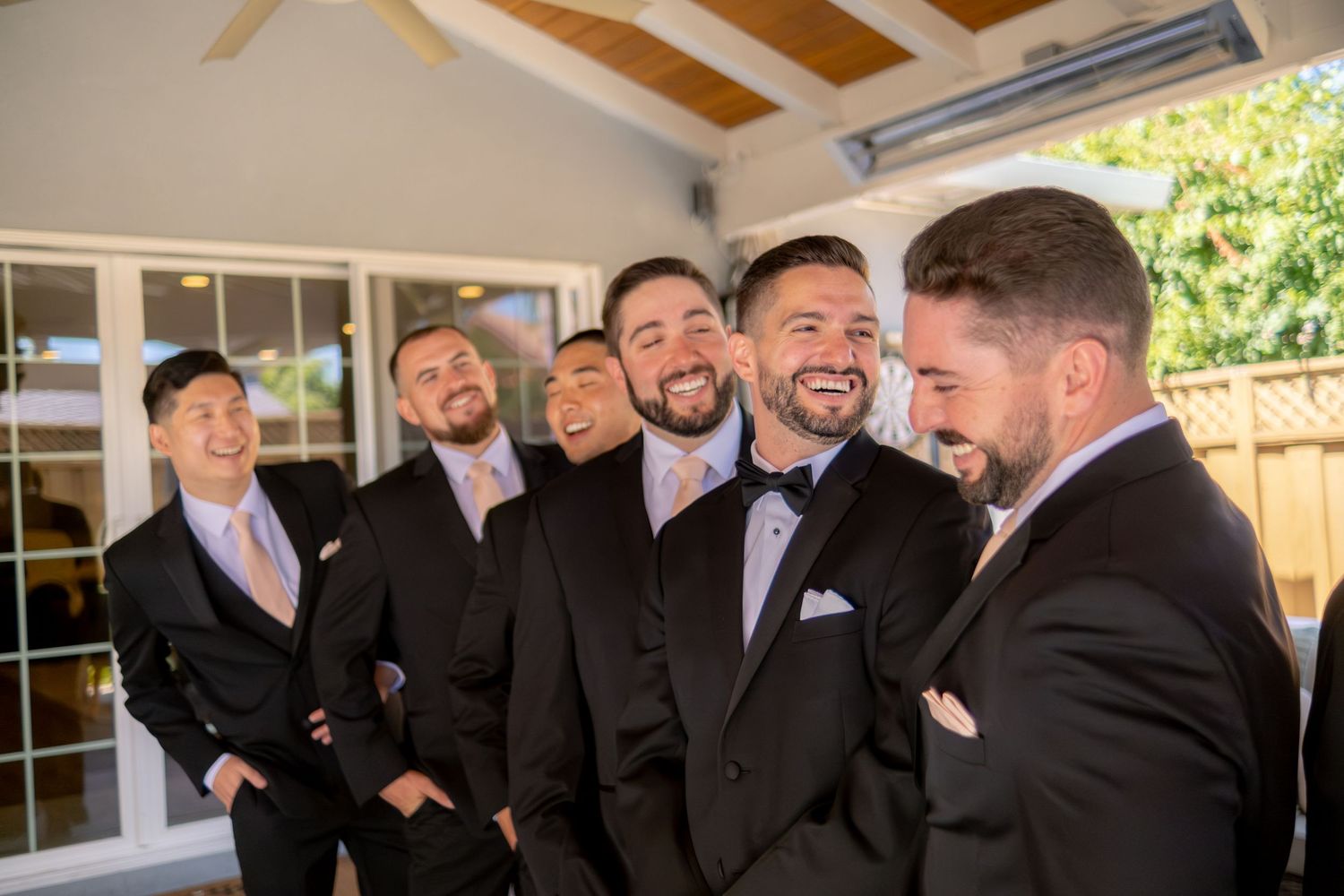 Groom and groomsmen laughing together in matching black tuxedos with bow ties before the wedding ceremony.