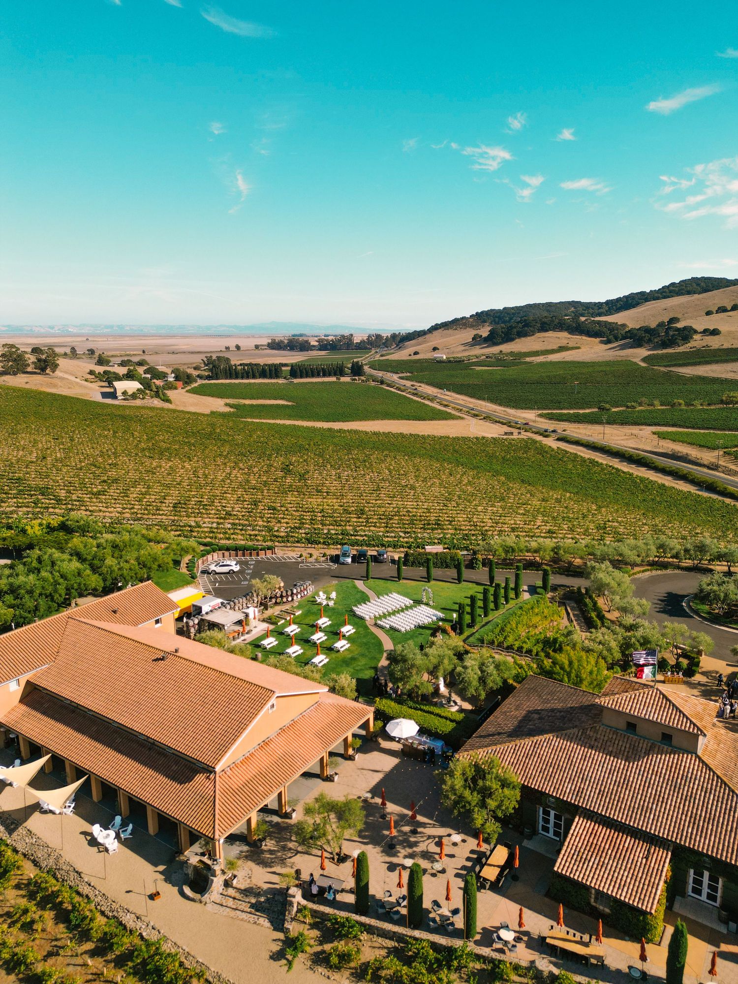 Aerial view of a California winery wedding venue surrounded by lush vineyards and rolling golden hills under a clear blue sky.