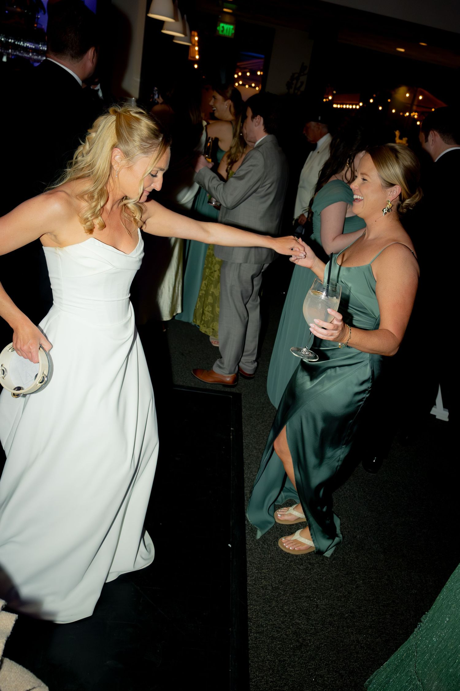 Bride in white strapless gown dances joyfully with bridesmaid in green satin dress at wedding reception.