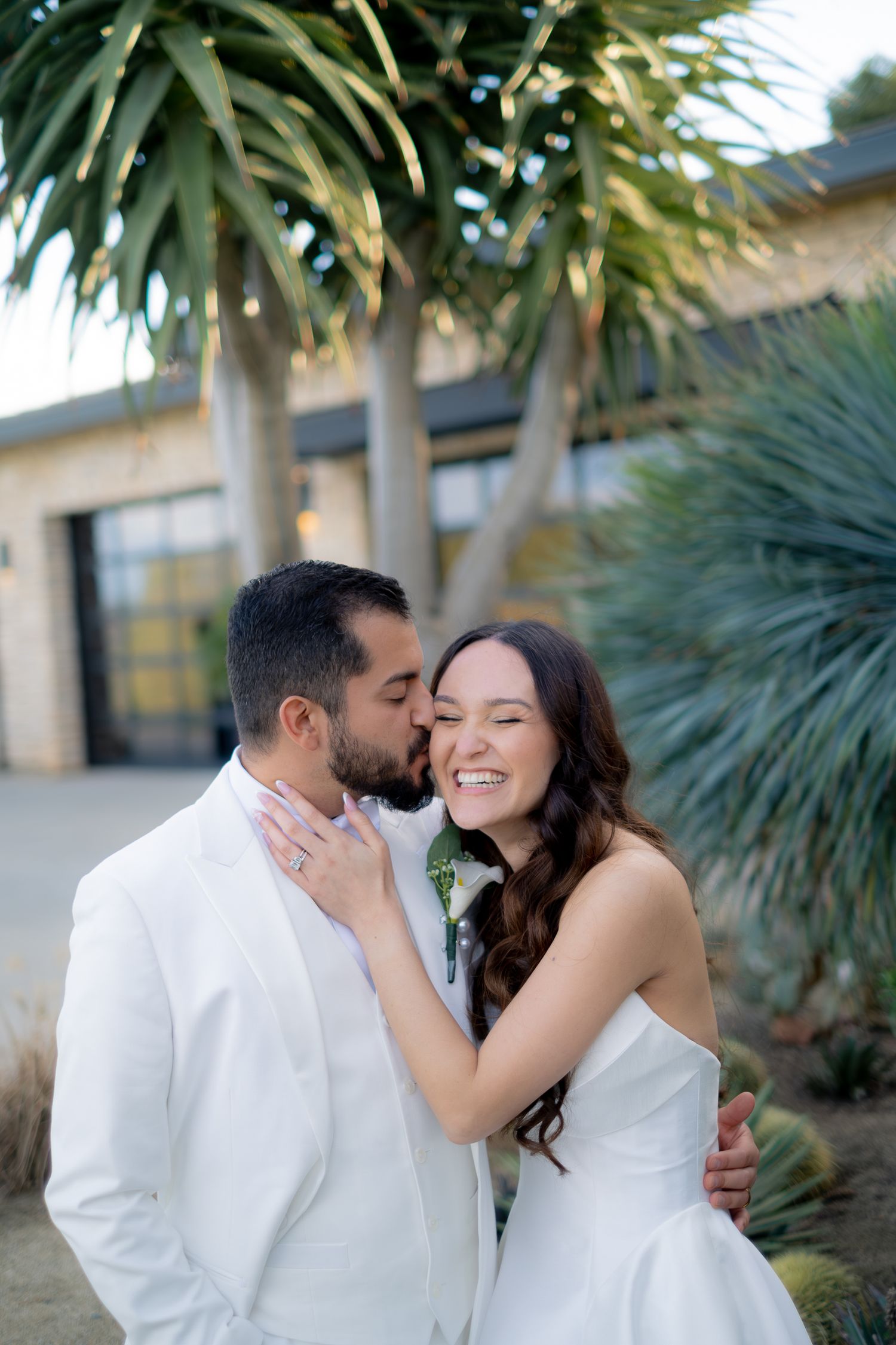 Groom in white suit kisses smiling bride on the cheek outdoors near tropical palm trees.