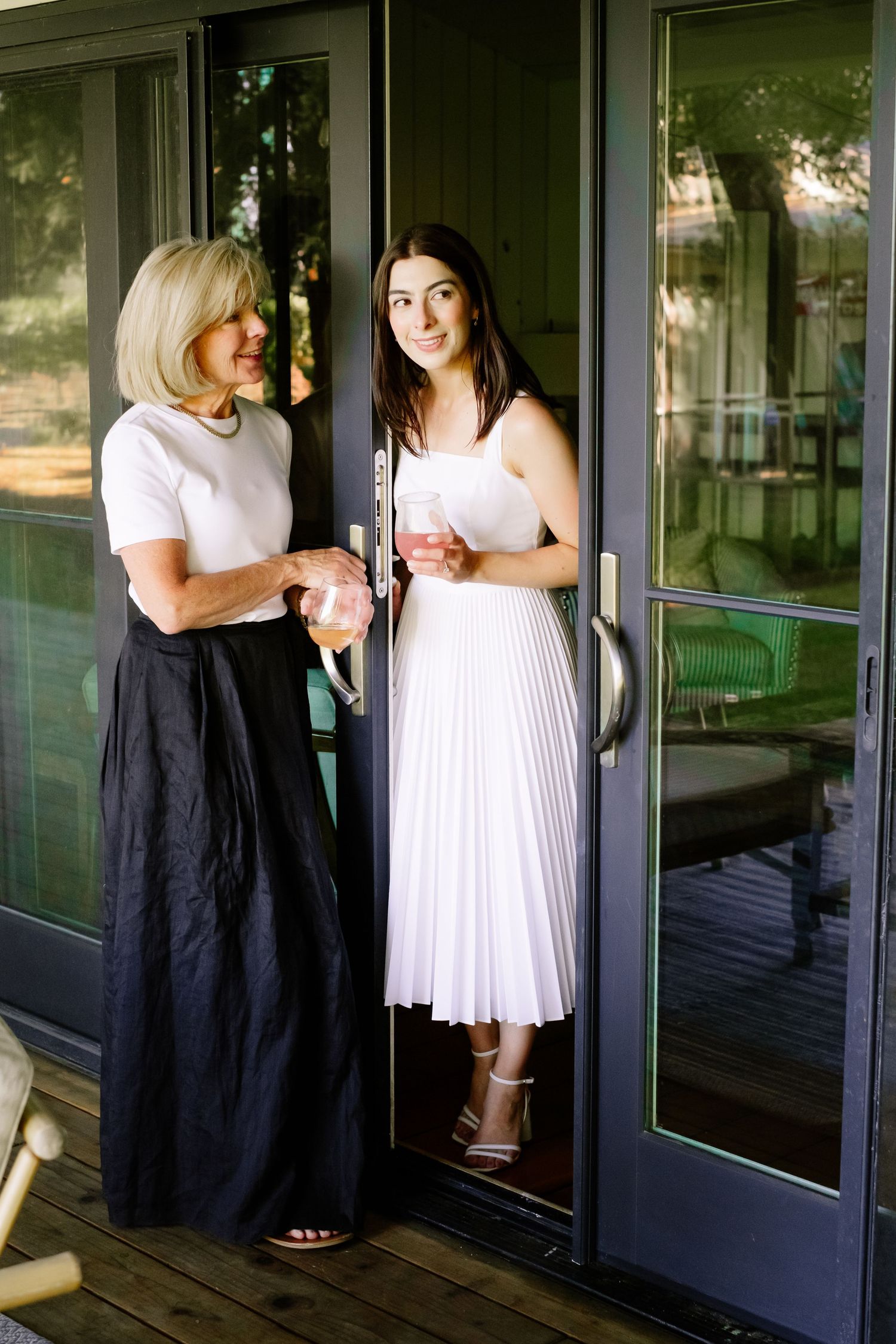 Blonde woman and brunette bride in white pleated dress holding drinks by glass doors at elegant venue.