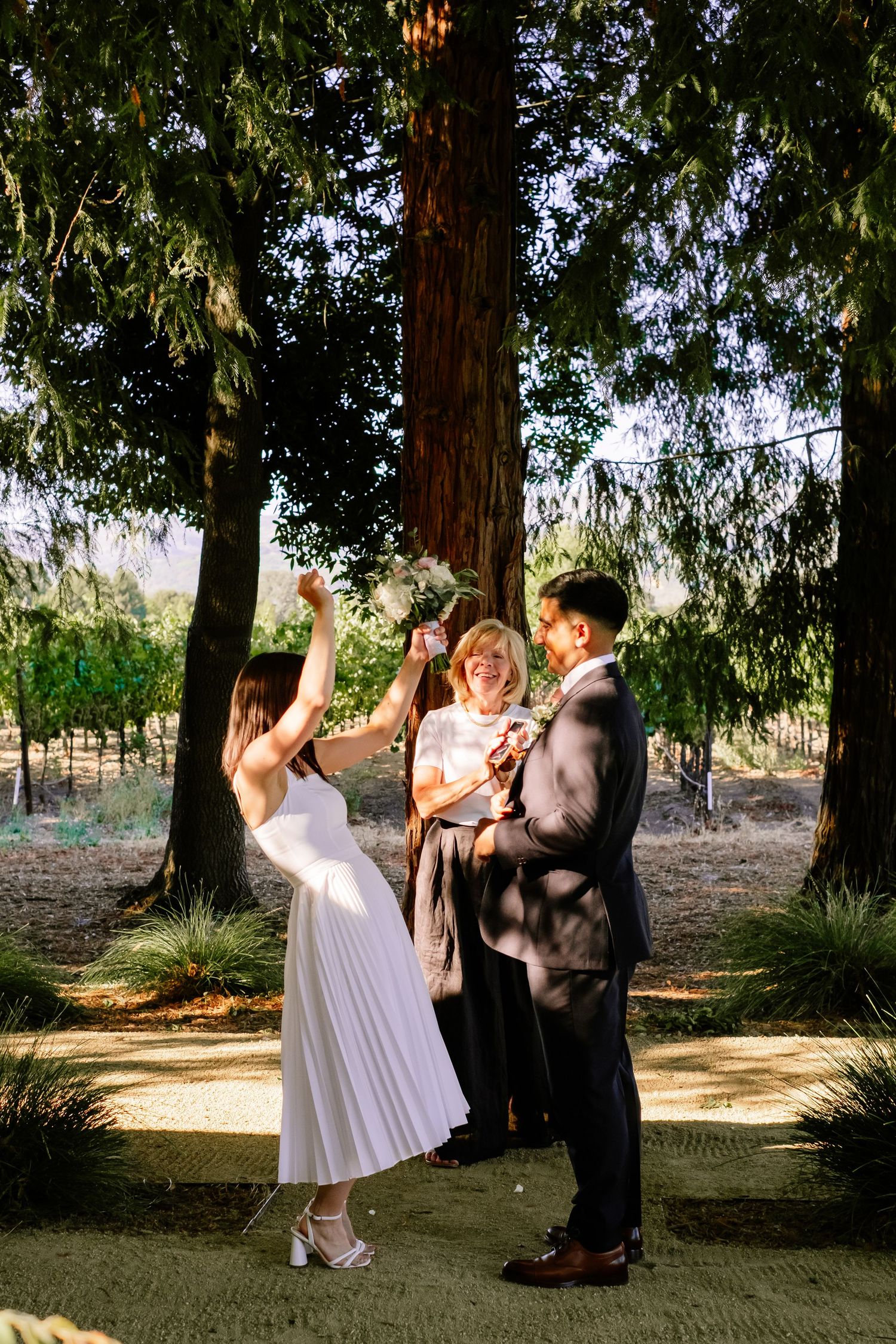 Bride raises hand joyfully during outdoor wedding ceremony under tall redwood trees with officiant and groom.