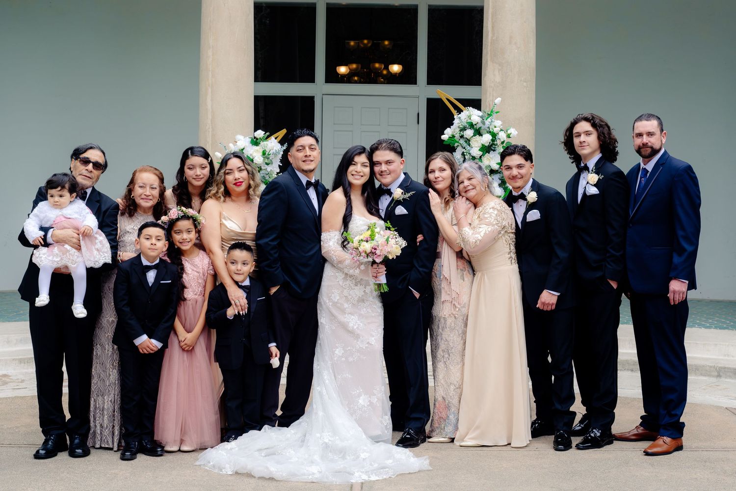 Wedding party poses together outside venue entrance with bride in white gown holding bouquet.