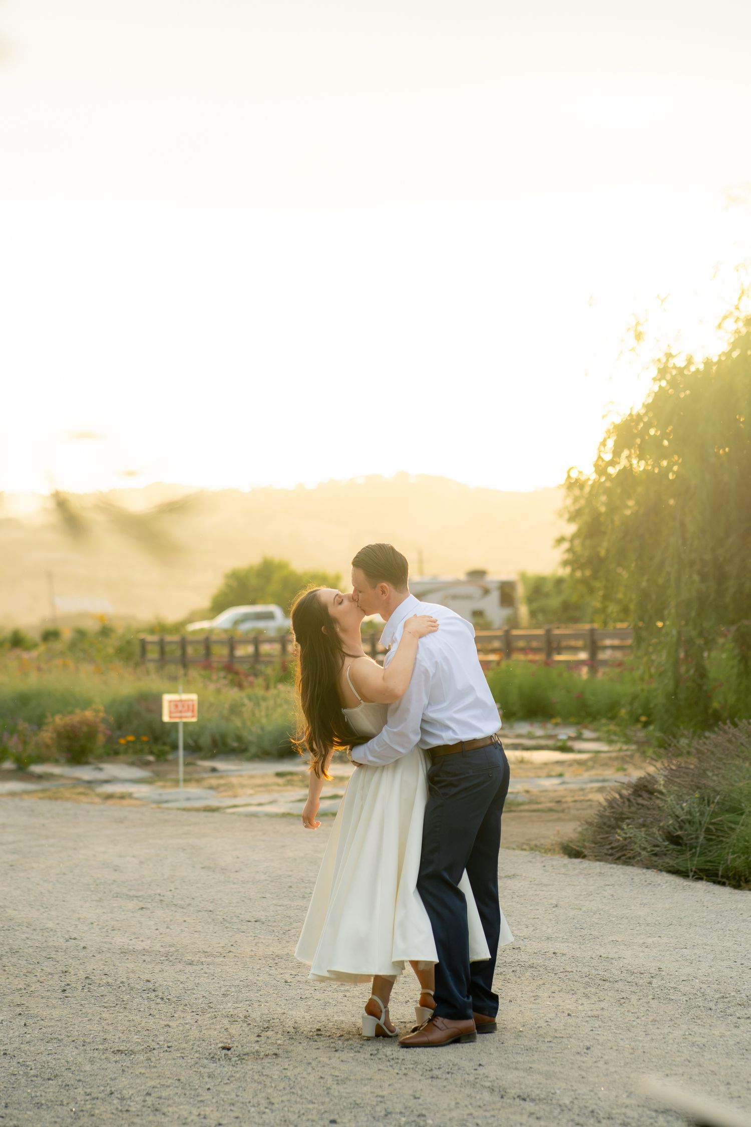 Couple sharing a romantic kiss at golden hour on a gravel path with rolling hills and warm sunset light behind them.