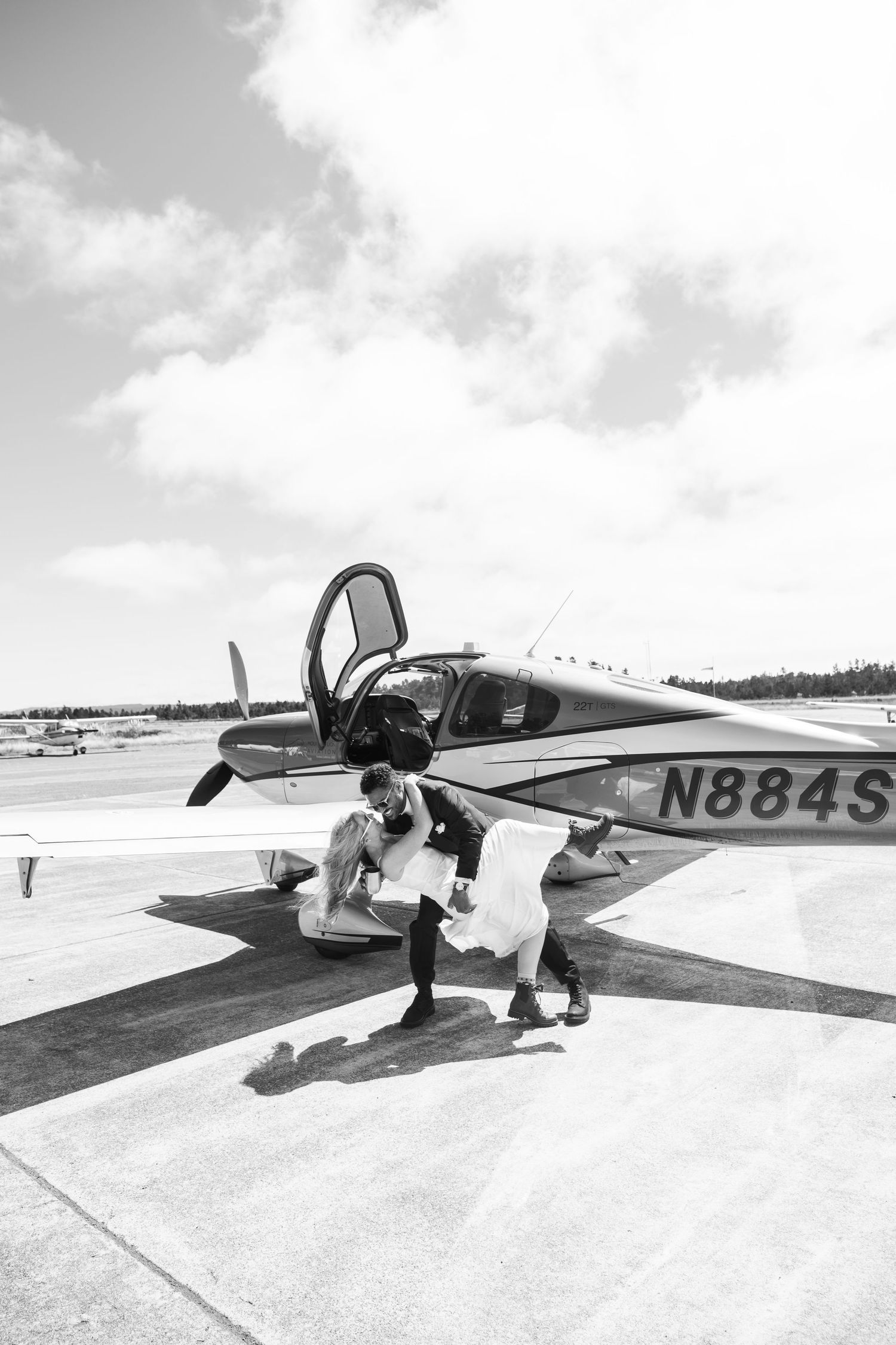 Black and white photo of a couple in wedding attire near a small propeller plane with tail number N884S on an airport tarmac.