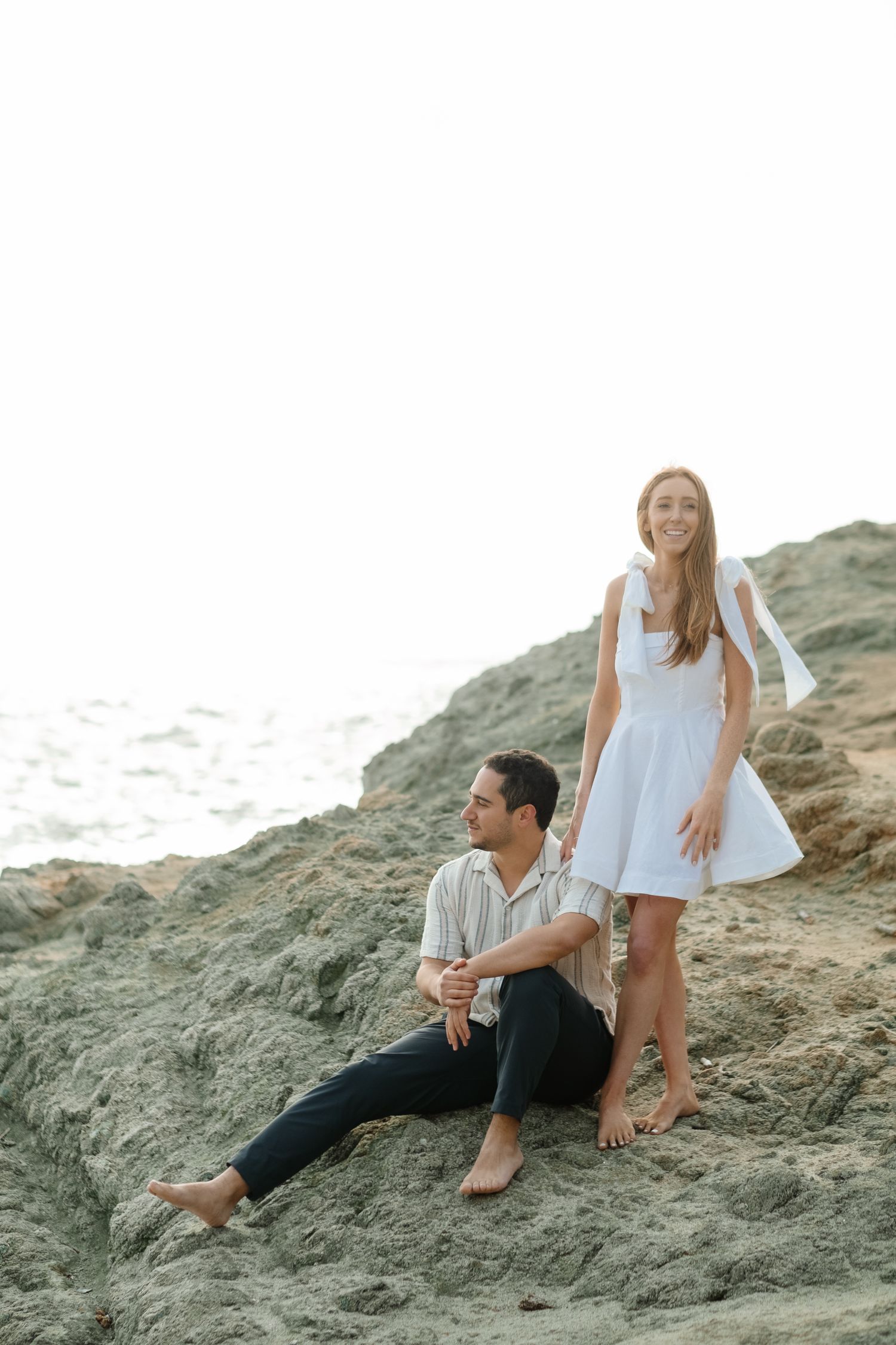 A couple sits on coastal rocks near the ocean, woman in white dress standing beside seated man.
