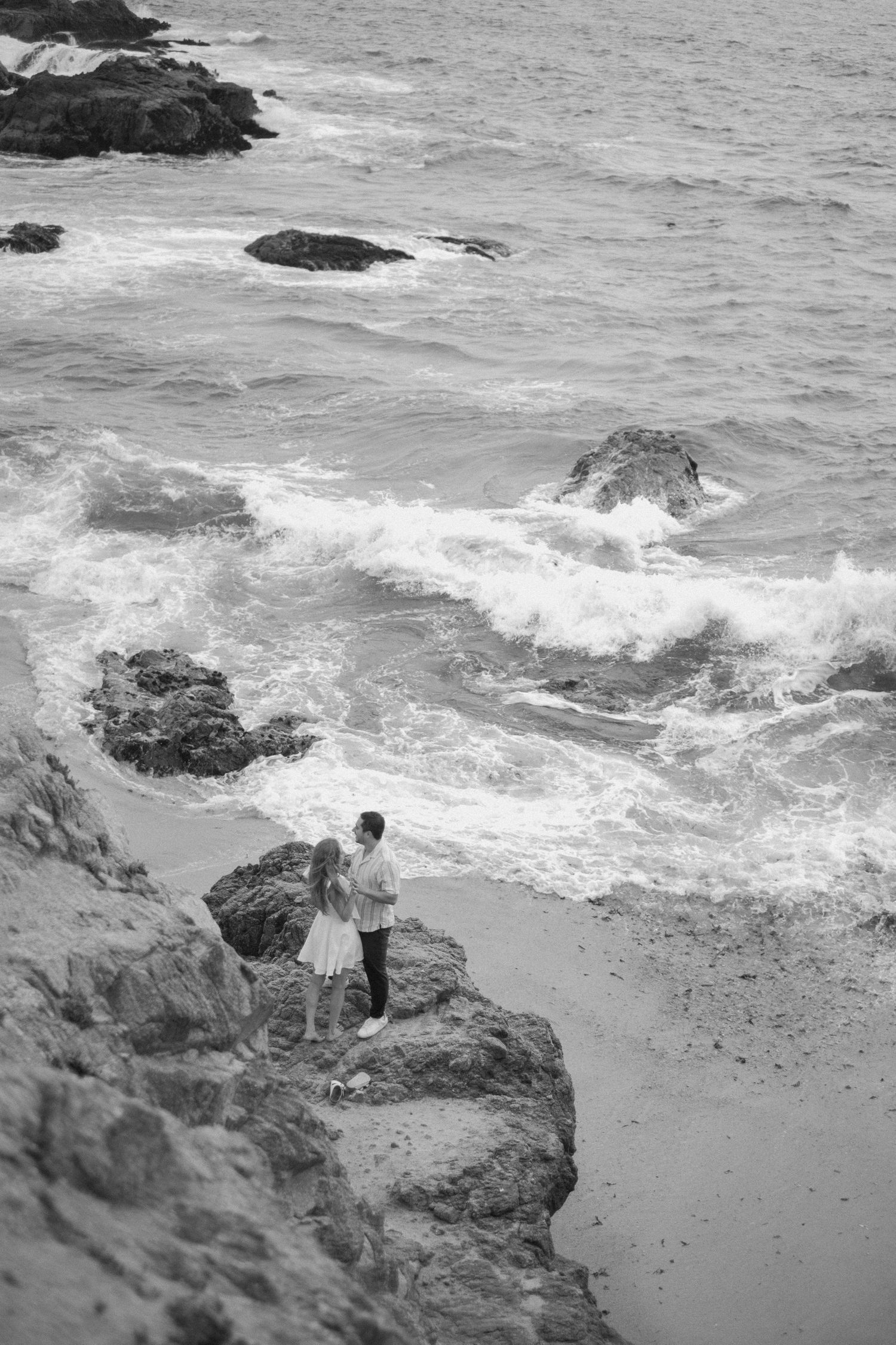 Black and white aerial view of couple standing on coastal rocks with crashing ocean waves below.