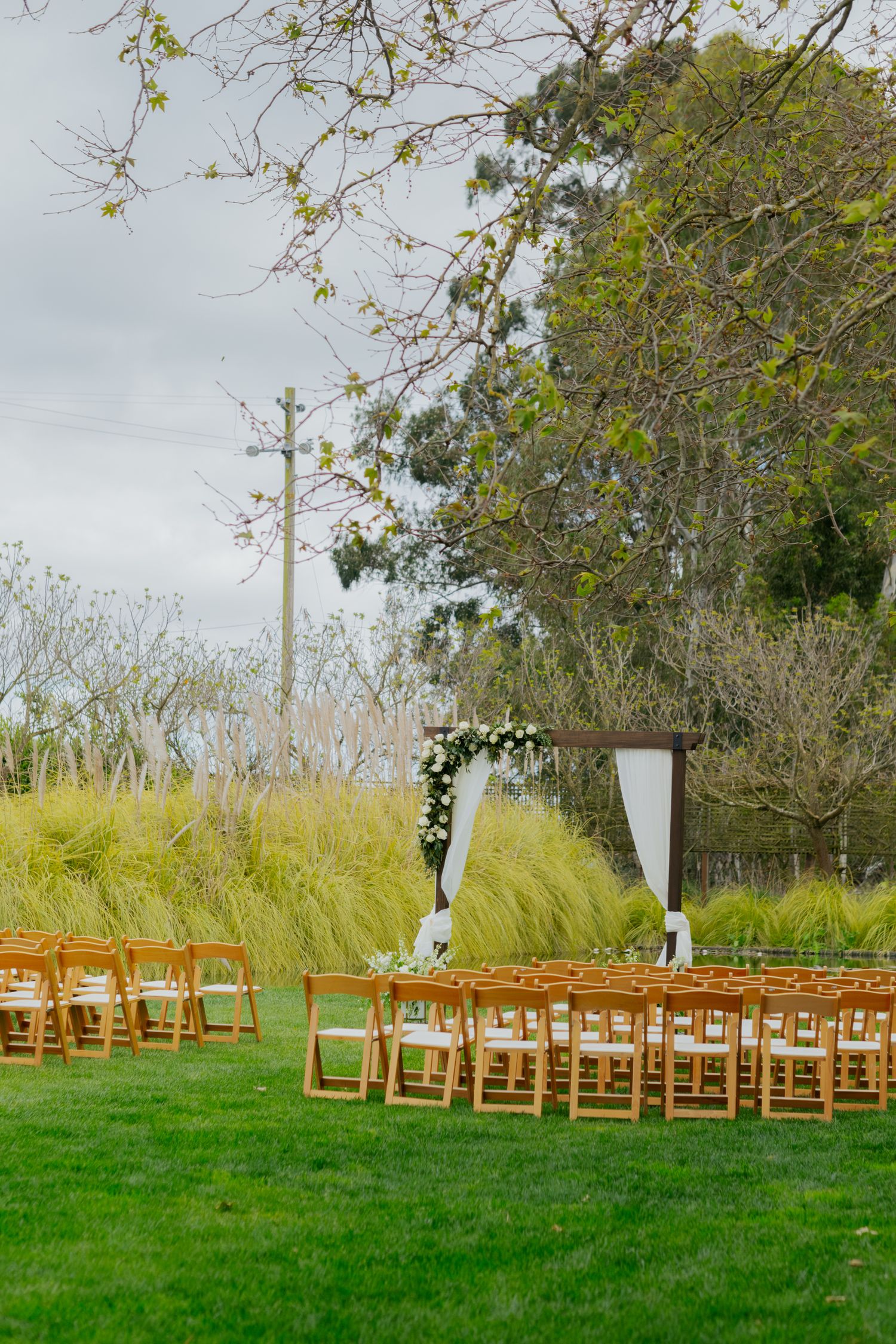 Outdoor garden wedding ceremony setup with wooden folding chairs, rustic wooden arch, and white floral arrangements on green lawn.