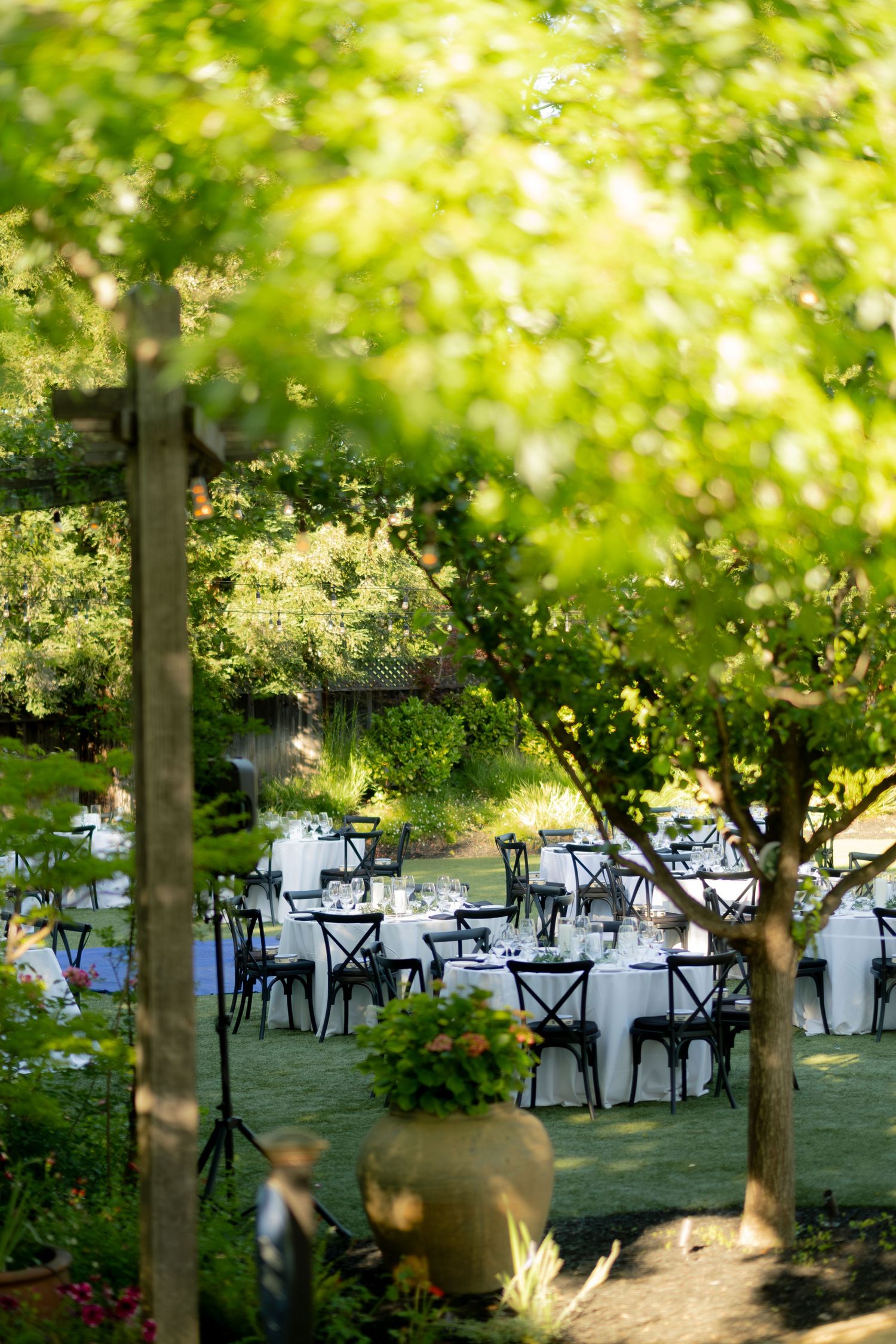 Elegant outdoor wedding reception tables with white linens set among lush green trees in garden.
