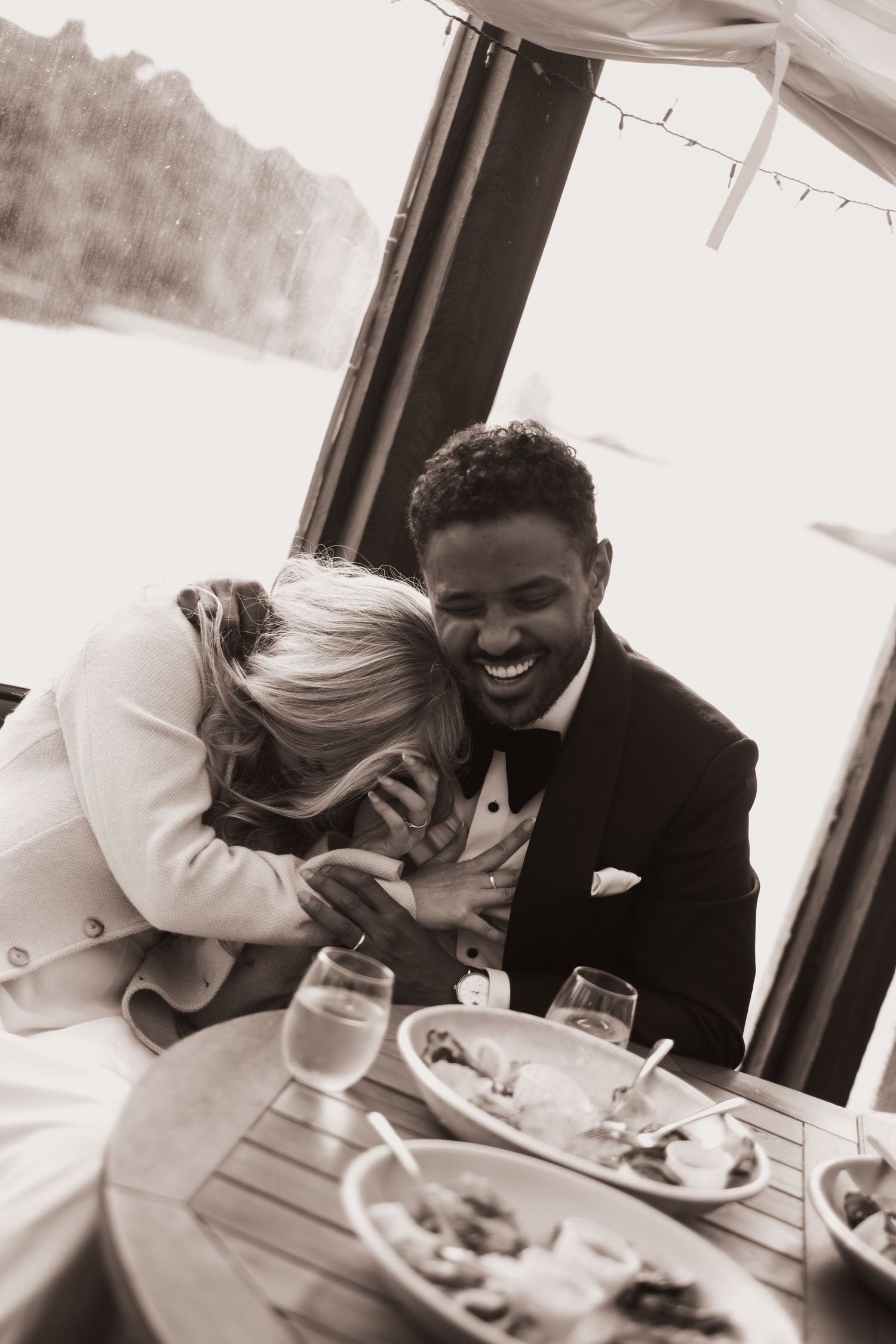 Black and white photo of laughing bride and groom sharing joyful moment at wedding reception.