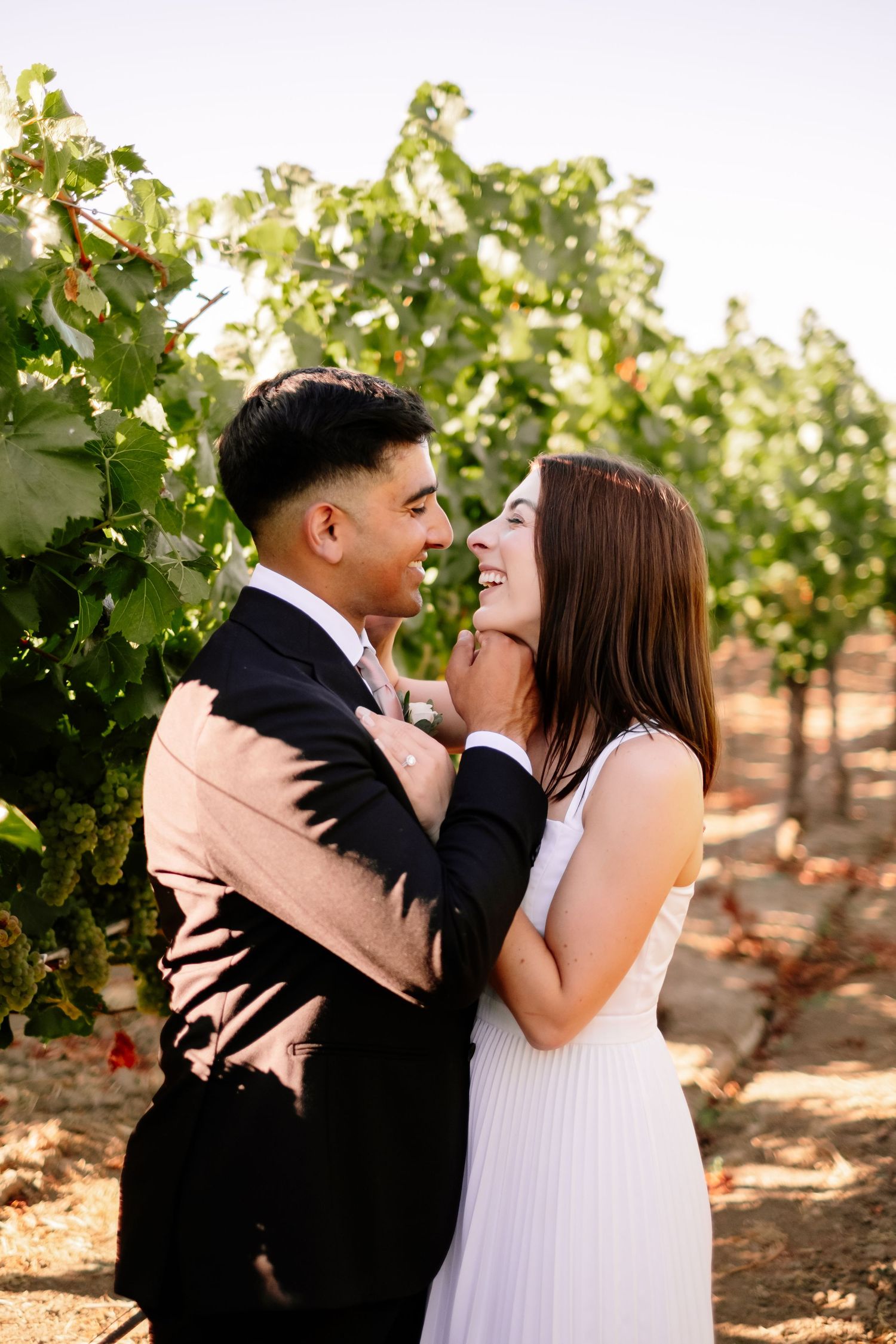 Smiling bride and groom share a romantic moment together in a sunny vineyard setting.