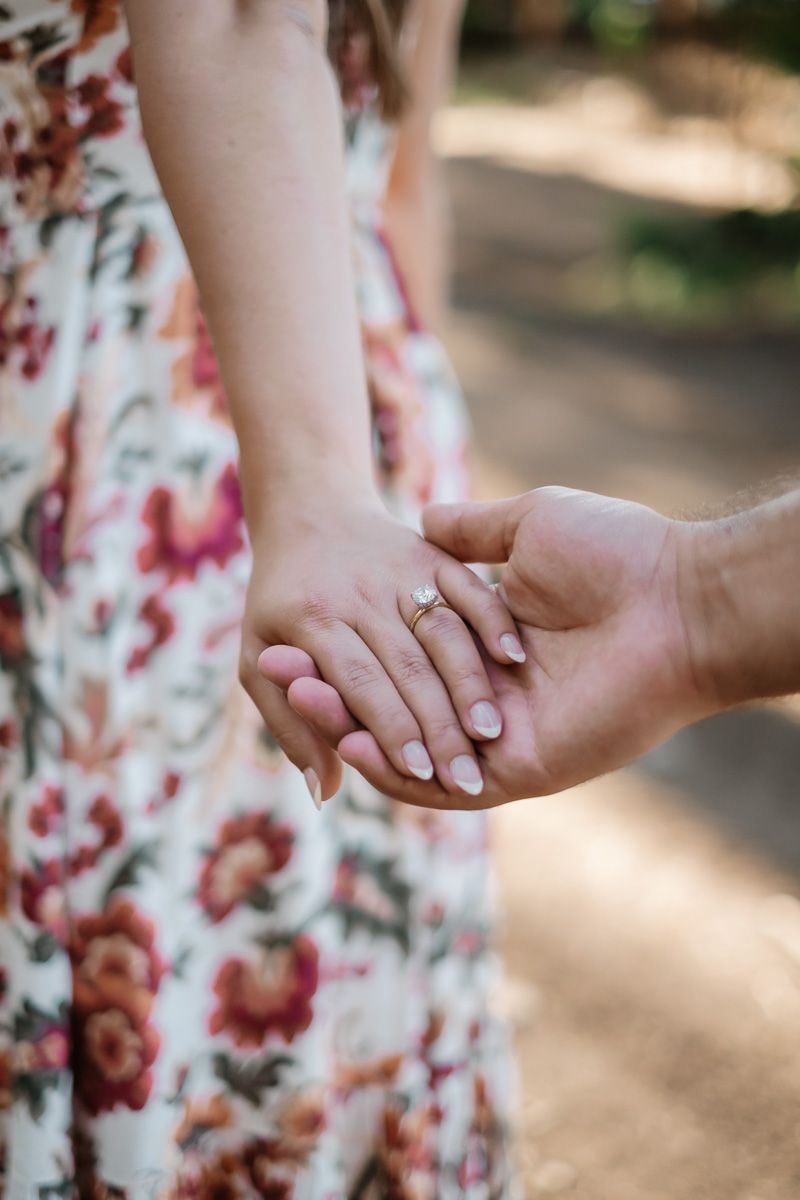 Couple holding hands outdoors, woman wearing floral dress and diamond engagement ring, romantic engagement photo.