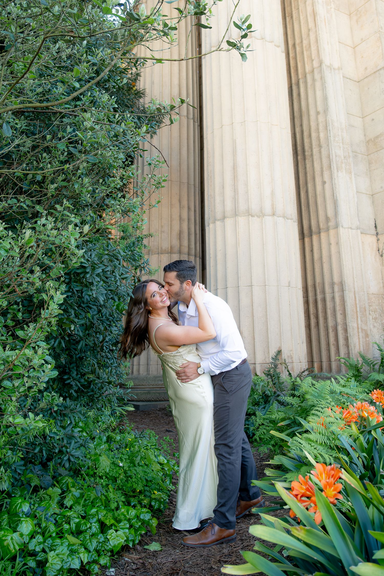 Couple embracing and kissing among lush greenery beside stone columns at the Palace of Fine Arts.