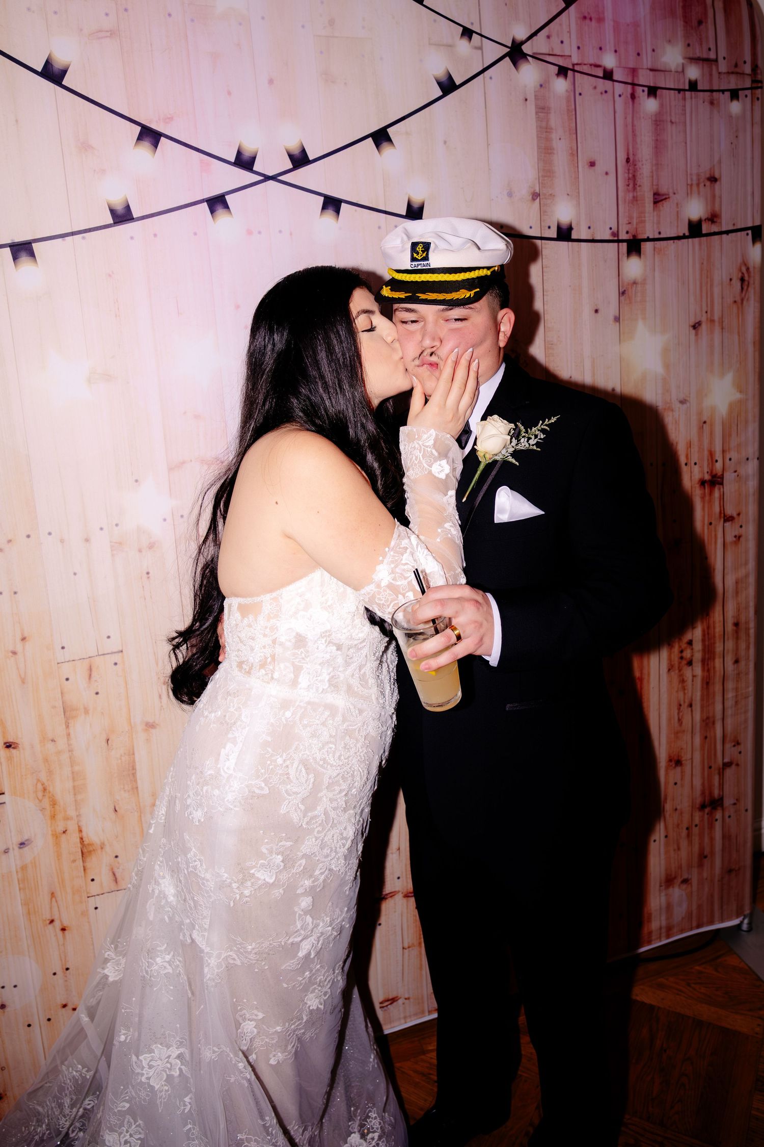 Bride kisses groom wearing captain hat at fun wedding photo booth backdrop.