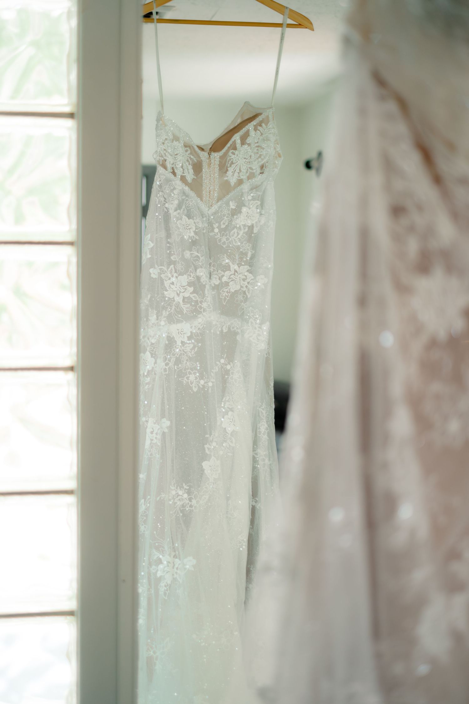 Elegant lace wedding dress hanging on a gold hanger near a bright window, featuring floral appliques and spaghetti straps.