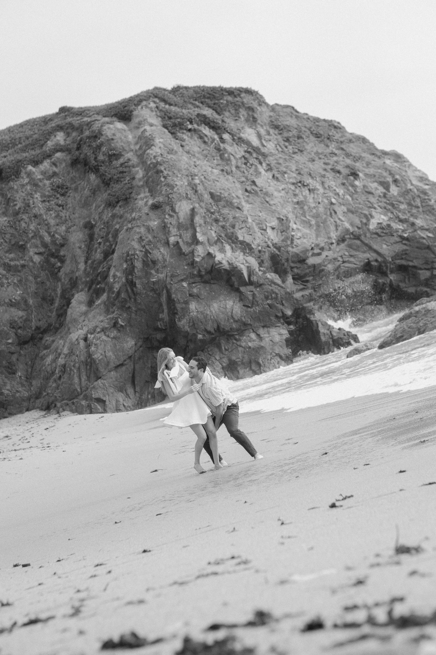 Black and white photo of a couple embracing on a sandy beach with rocky cliffs behind them.