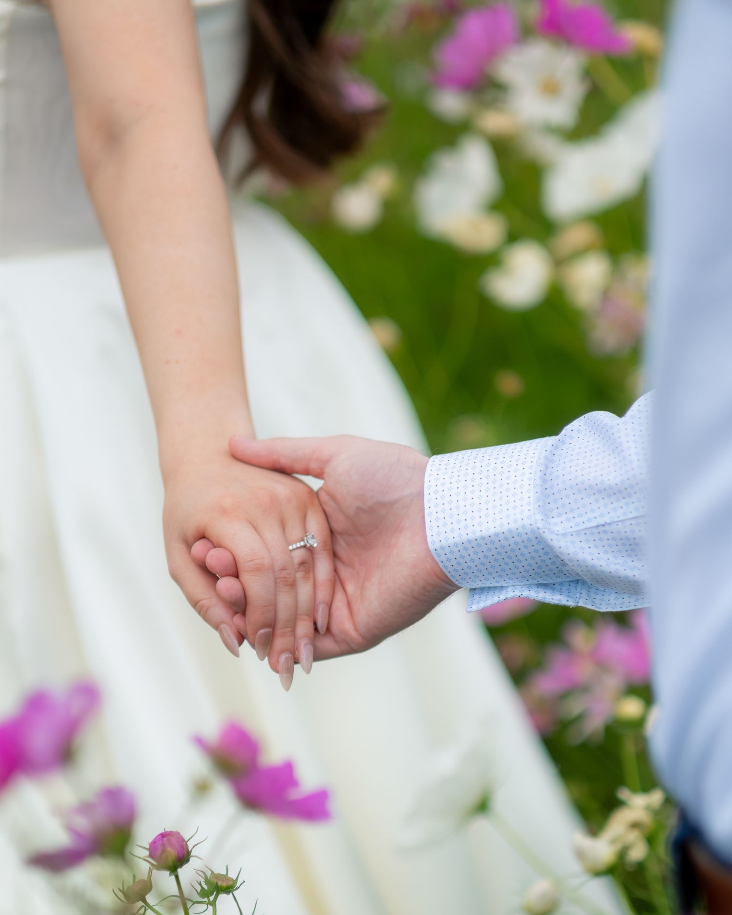 Close-up of engaged couple holding hands with diamond ring in flower field.