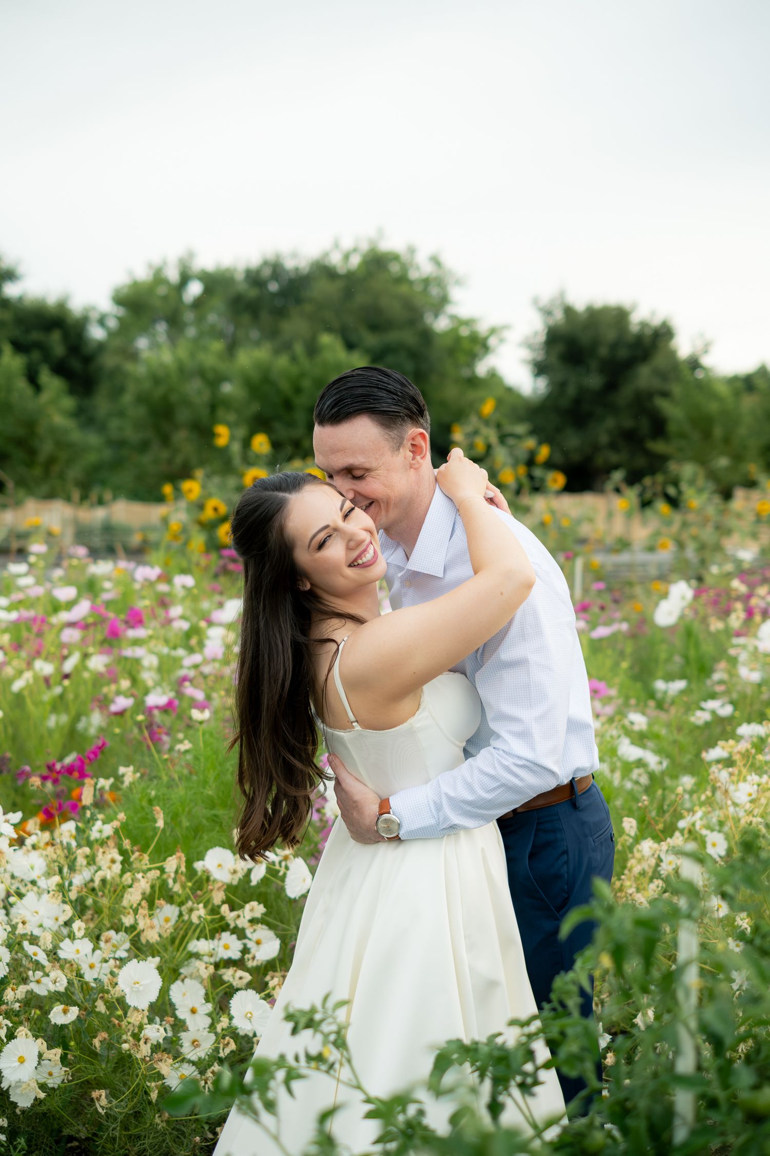 Couple embracing in a colorful wildflower garden, woman in white dress smiling as man kisses her cheek.