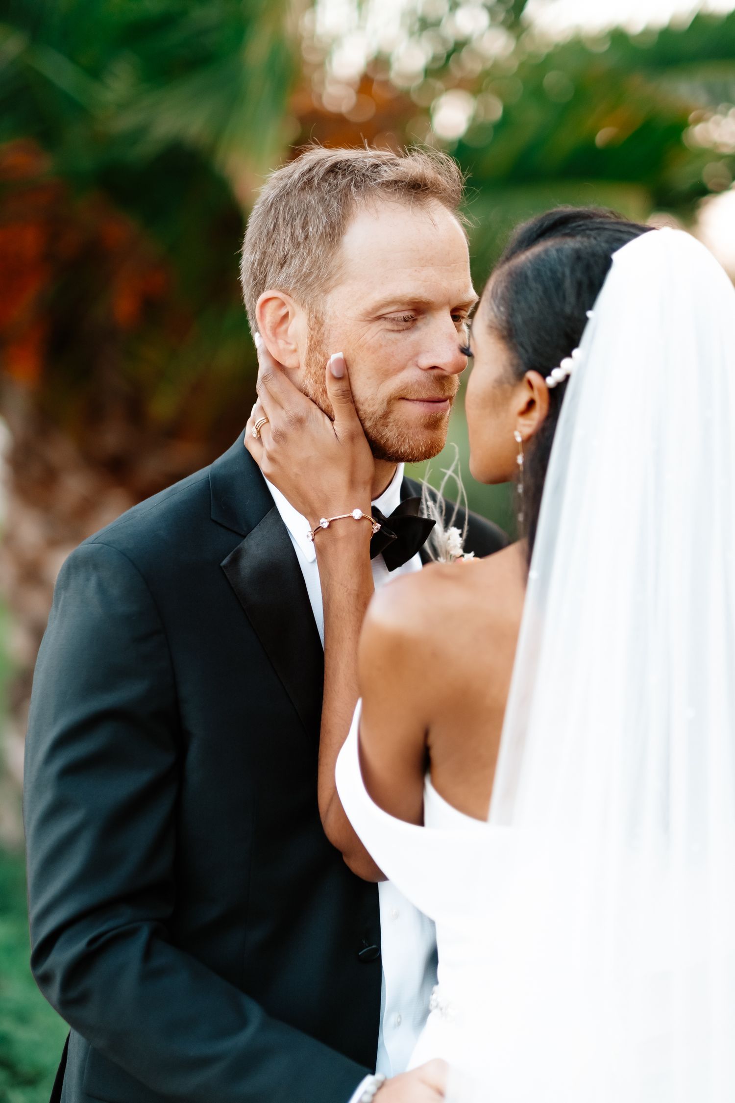 Groom in black tuxedo gazes tenderly as bride in white veil gently cups his face during outdoor wedding portraits.