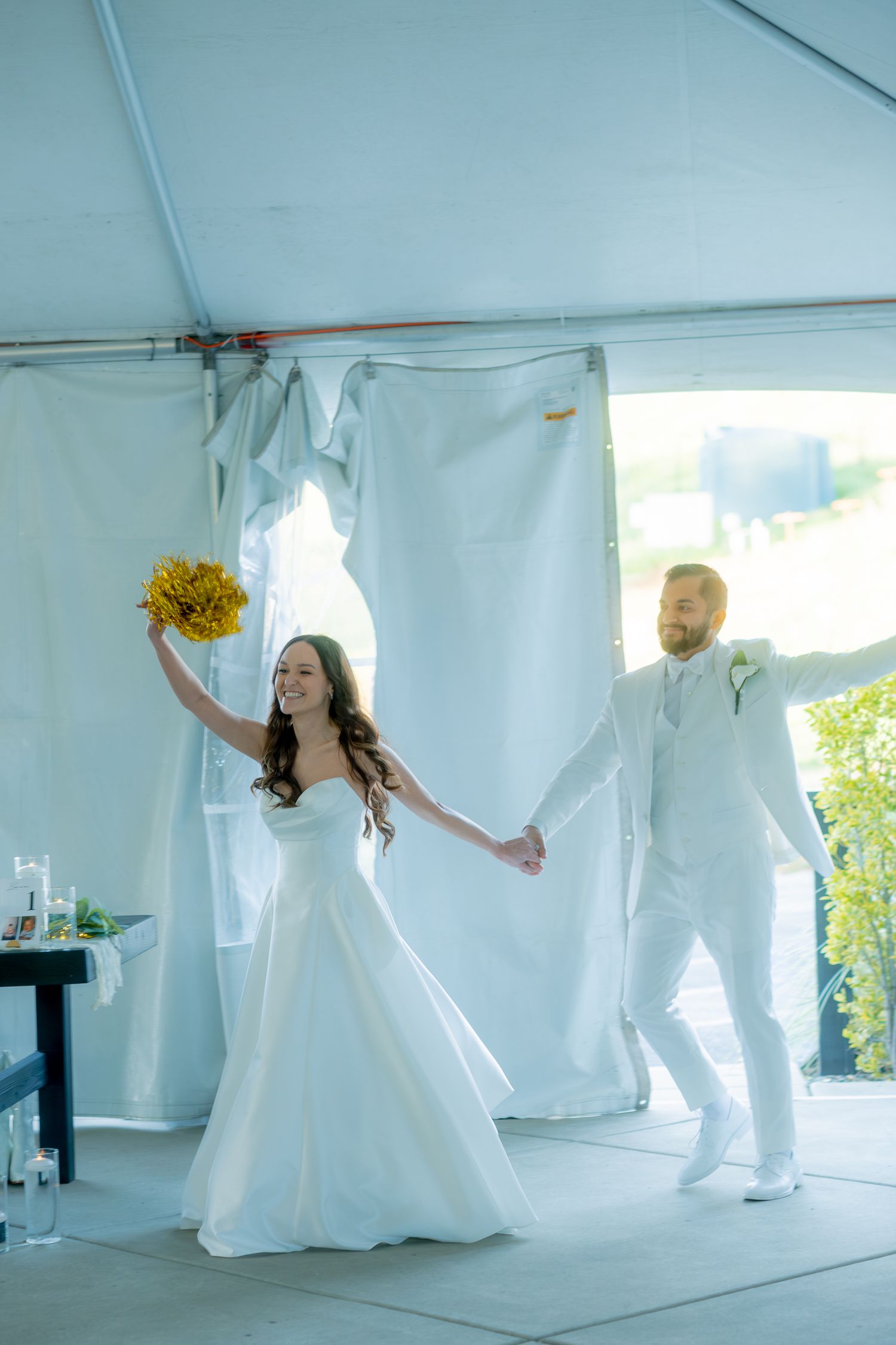 Joyful bride and groom make grand entrance under white tent, bride raising yellow bouquet, both dressed in white wedding attire.