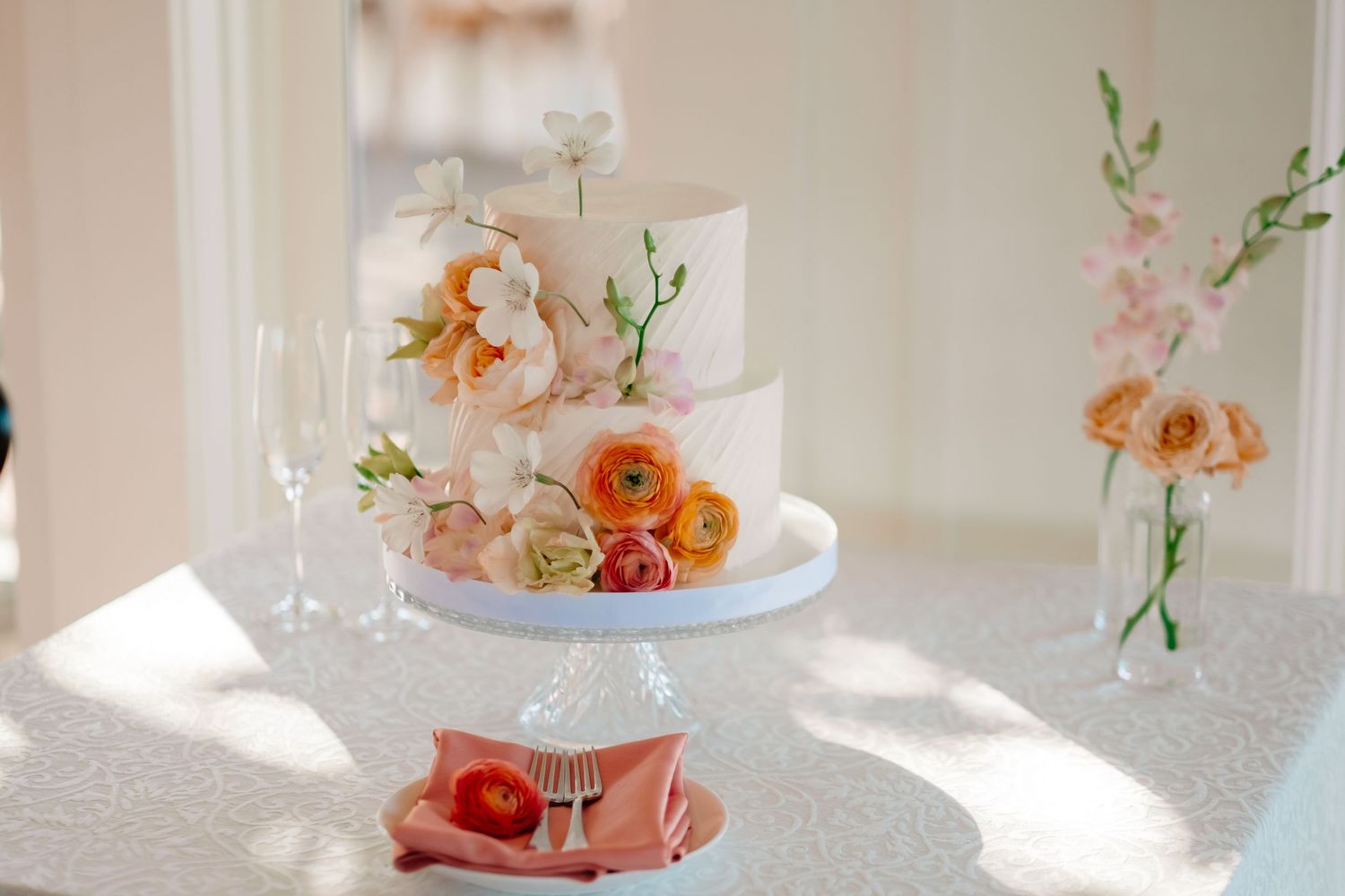 Elegant two-tier white wedding cake adorned with colorful peony and ranunculus flowers on a decorative cake stand.