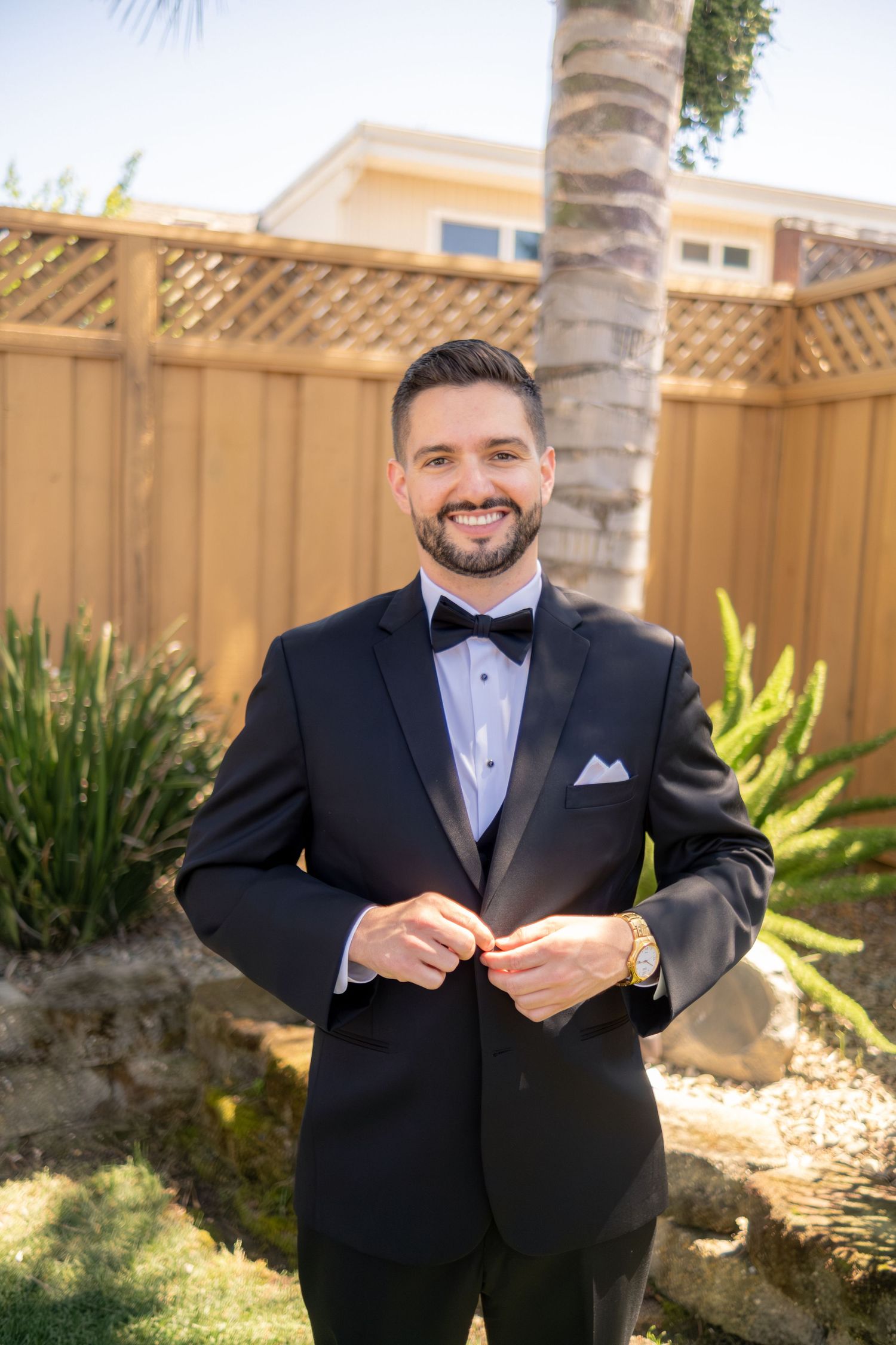 Handsome groom in black tuxedo with bow tie buttoning jacket while smiling in sunny backyard garden.