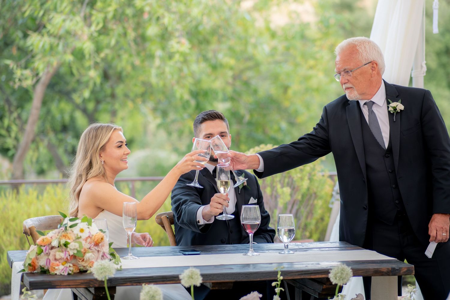 Bride and groom toasting champagne with older gentleman during outdoor wedding reception at sweetheart table.
