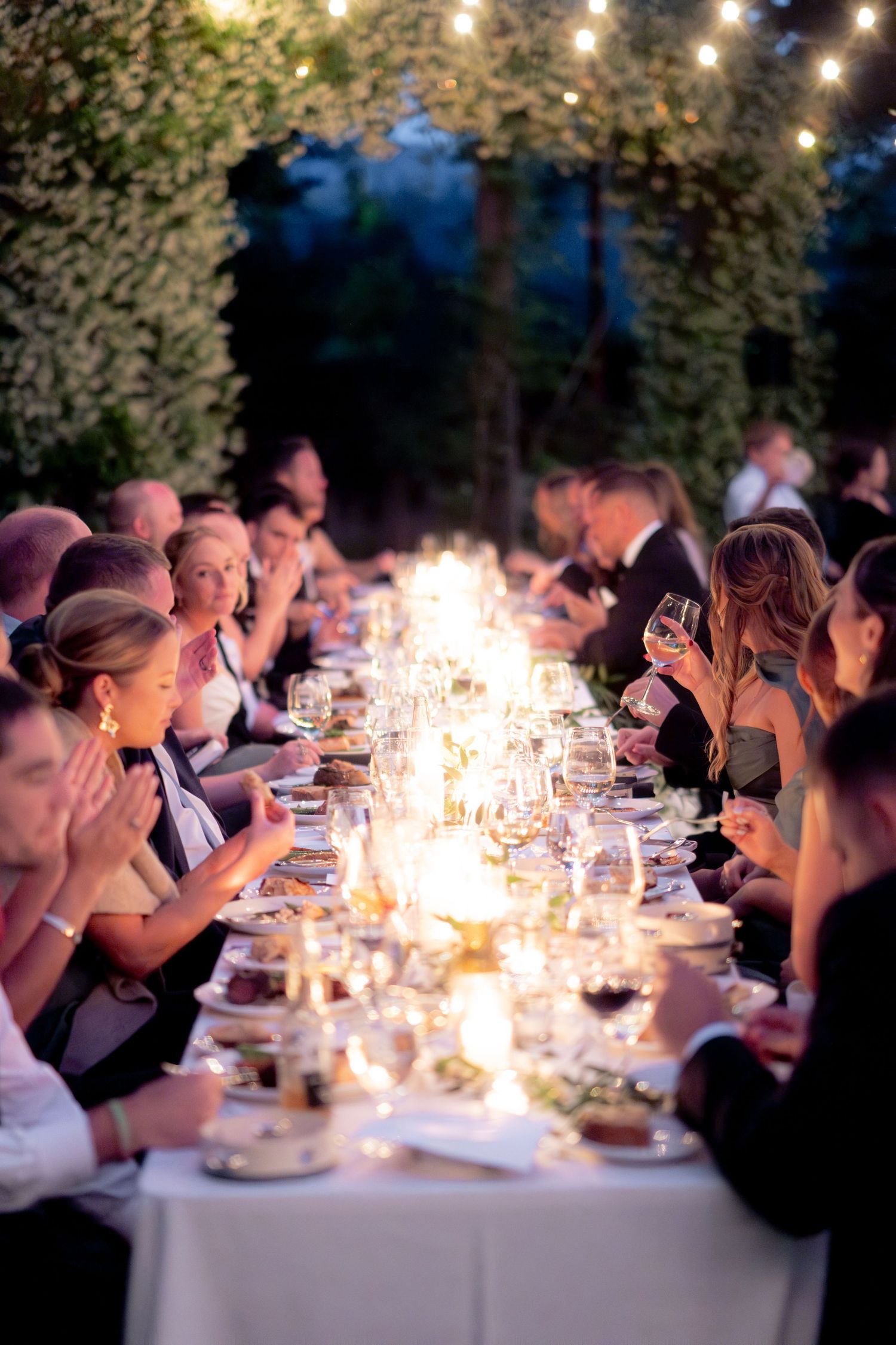 Elegant outdoor wedding reception dinner with long candlelit table, string lights, and guests enjoying wine.