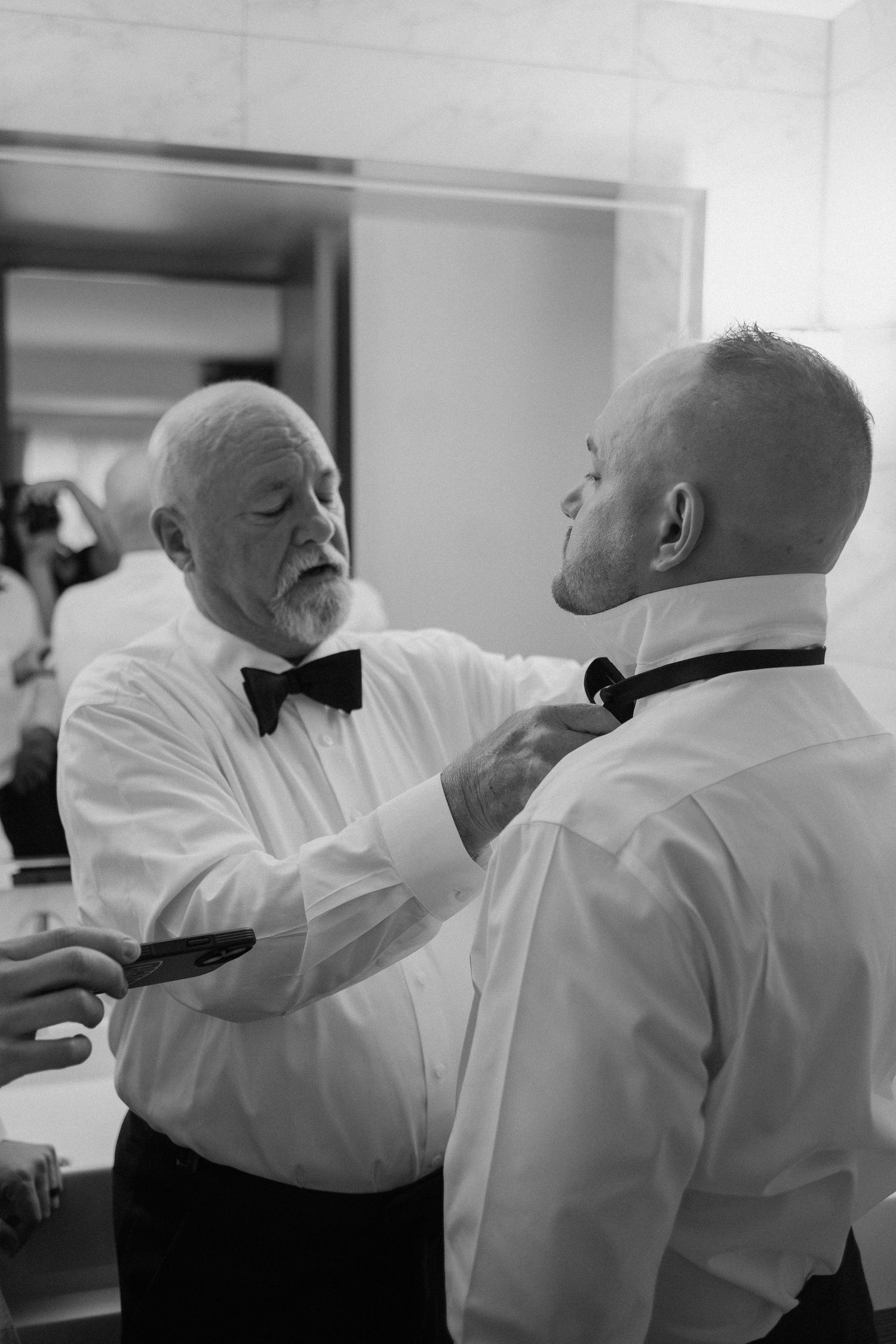 Black and white photo of older gentleman carefully adjusting grooms bow tie before wedding ceremony.