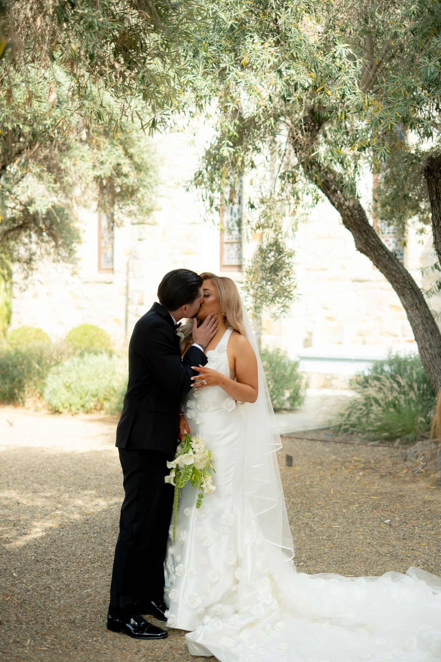 Bride and groom sharing romantic kiss under olive trees outside stone church.