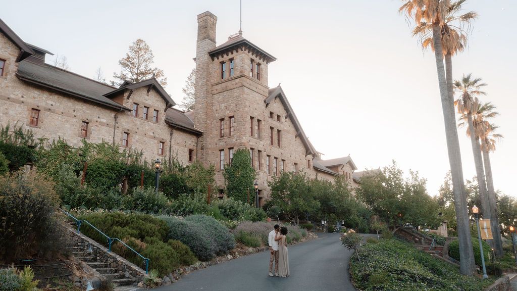 Couple walks along a path outside a historic stone castle building surrounded by lush greenery and tall palm trees.
