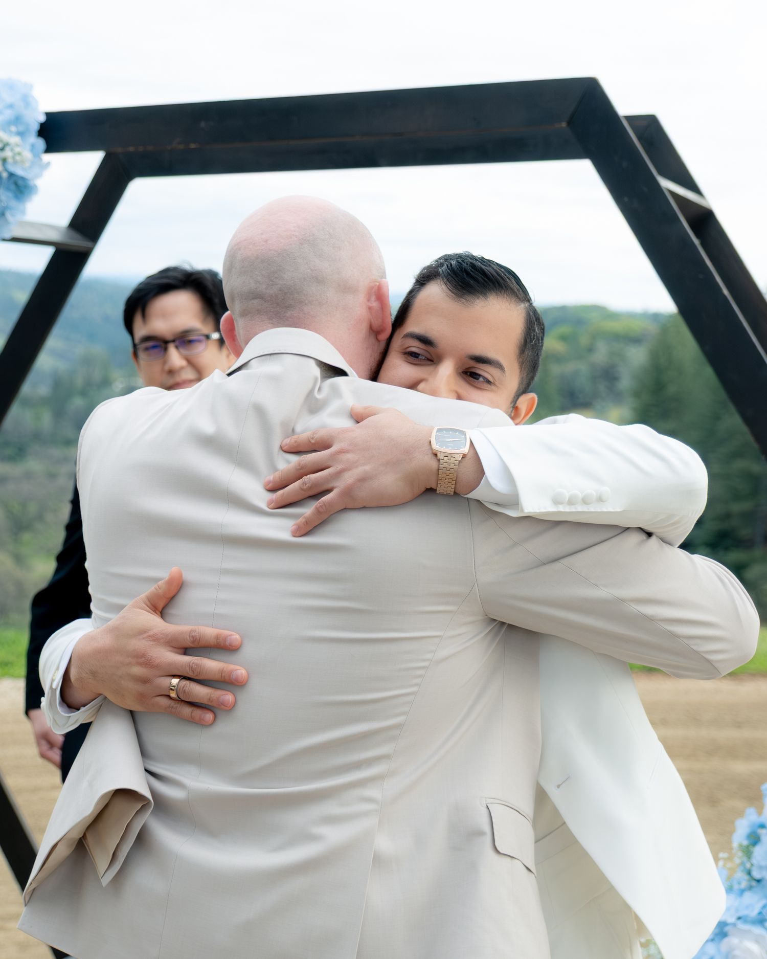 Groom in white suit embraces groomsman in tan suit during outdoor wedding ceremony.