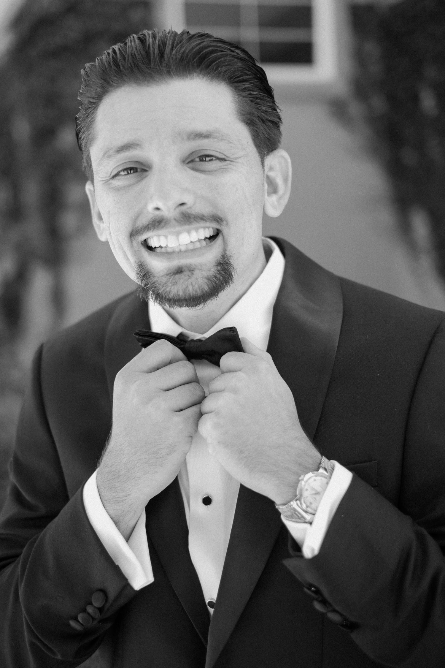 Dapper groom in black tuxedo adjusting his bow tie with a confident smile on his wedding day.