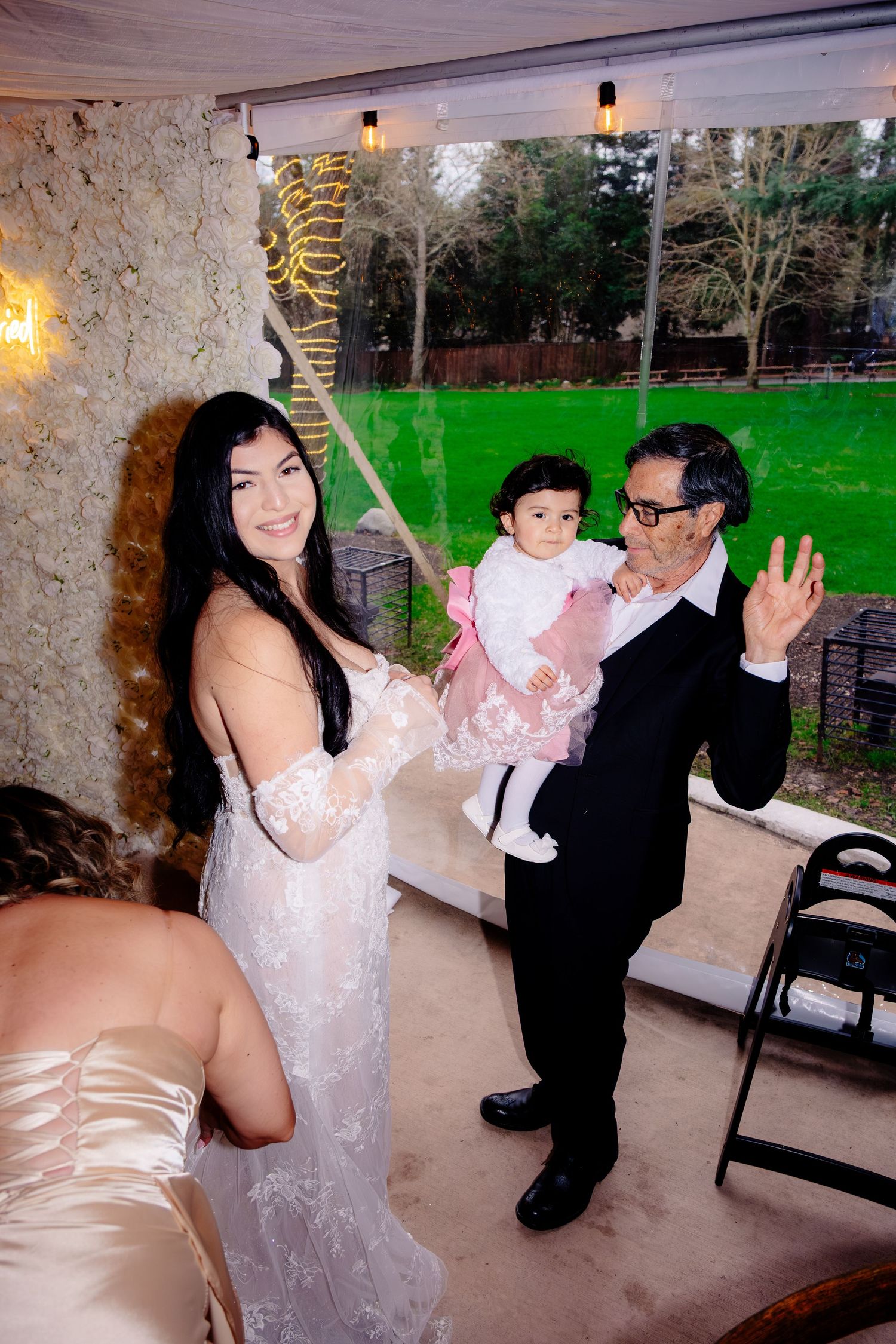 Bride in lace gown smiles with older man holding baby girl at elegant wedding reception venue.