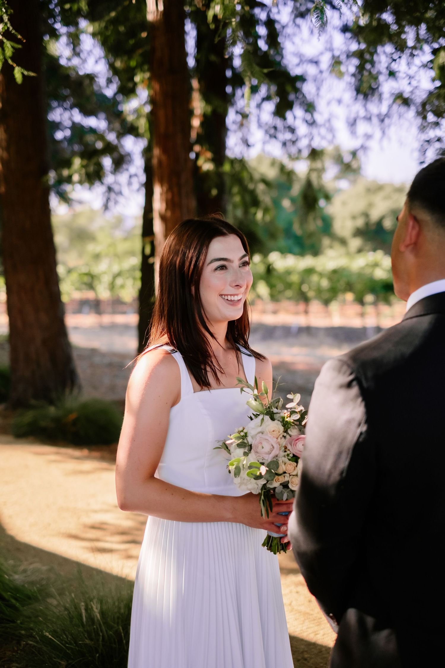 Smiling bride holding pink bouquet faces groom under tall trees in golden sunlight.