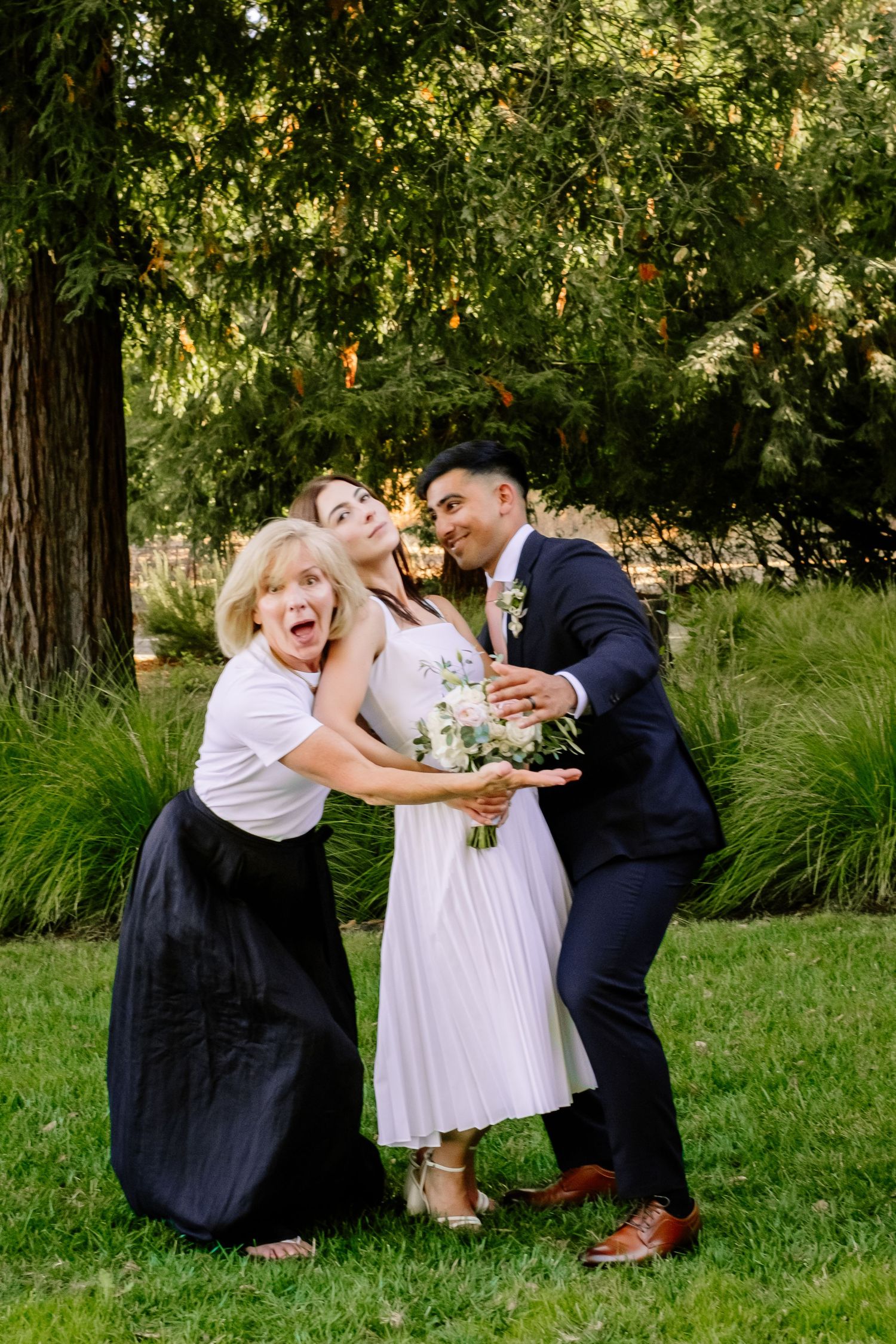 Joyful bride in white dress with groom in navy suit and excited blonde woman posing outdoors.